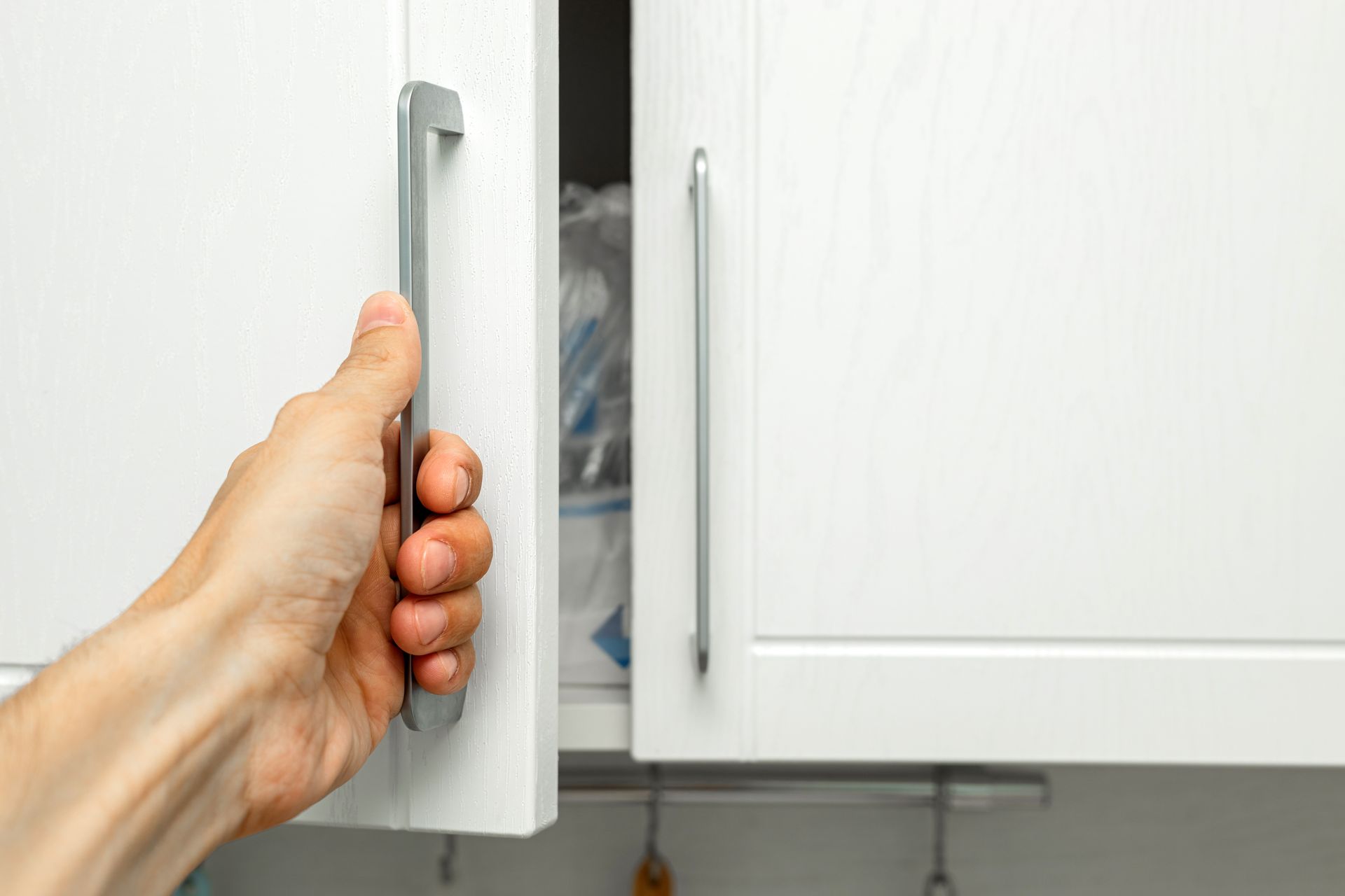 A hand pulls open a white kitchen cabinet door with a silver handle.