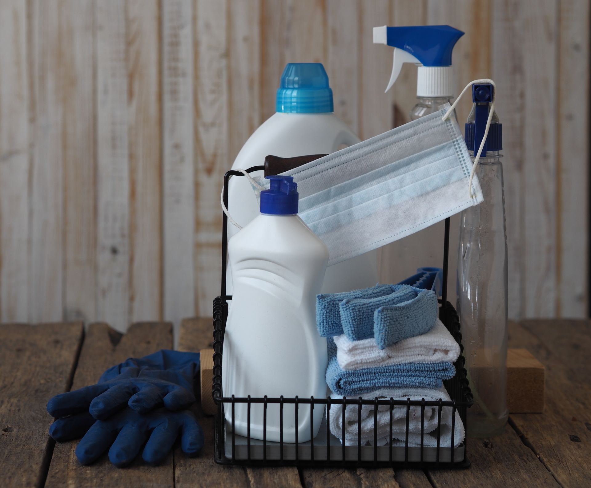 A cleaning caddy containing cleaning supplies, folded cloths, and a surgical mask sits on a wooden surface with blue gloves.