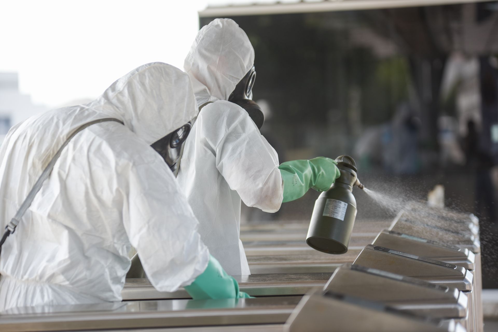 Two people in full white protective gear and green gloves spray disinfectant on a row of transit turnstiles.
