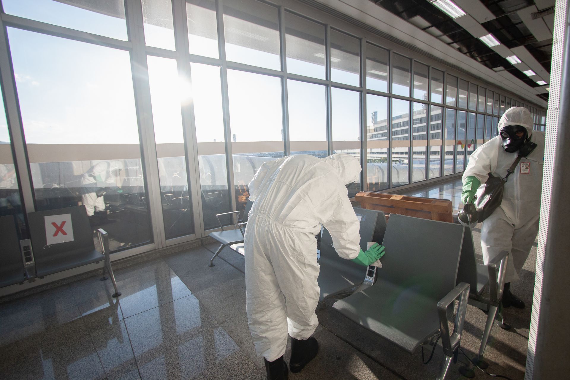 Two workers in white hazmat suits and masks disinfect airport seating near a large window.