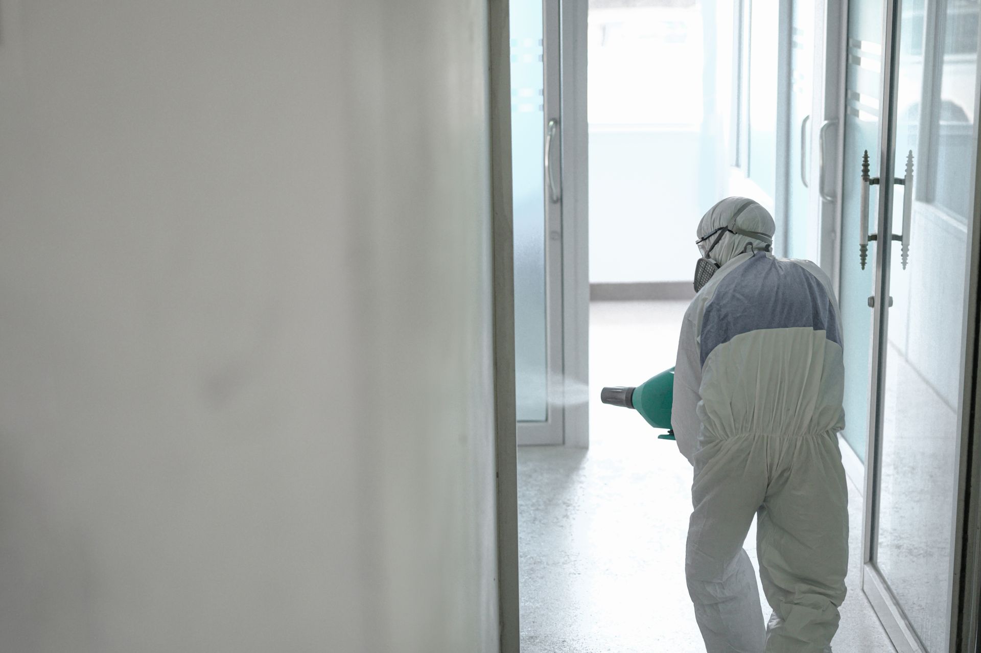 A person in white protective gear sprays disinfectant in a bright, modern corridor.