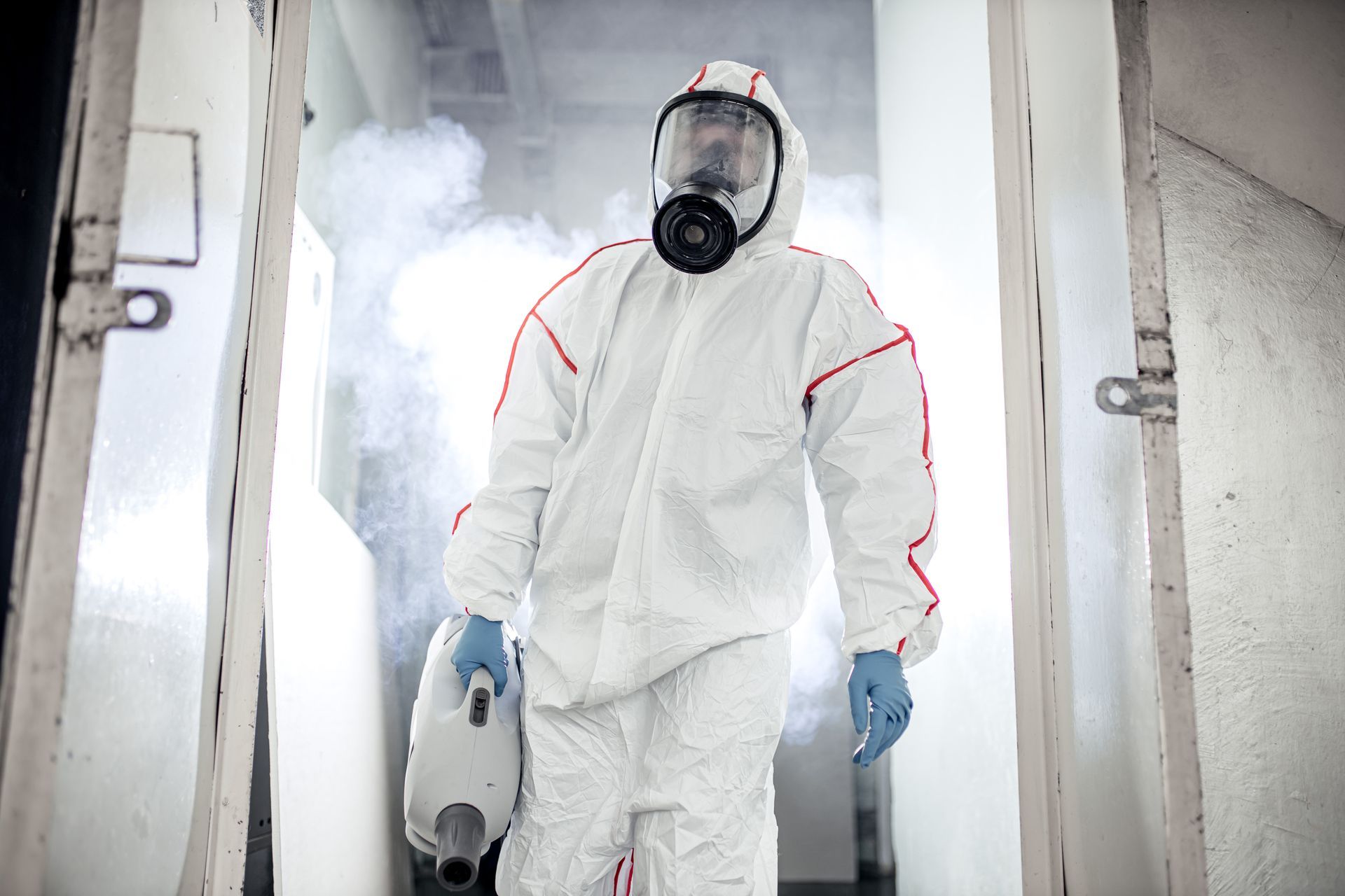 A person in a white hazmat suit and respirator mask walks through a doorway filled with mist while holding a fogger.