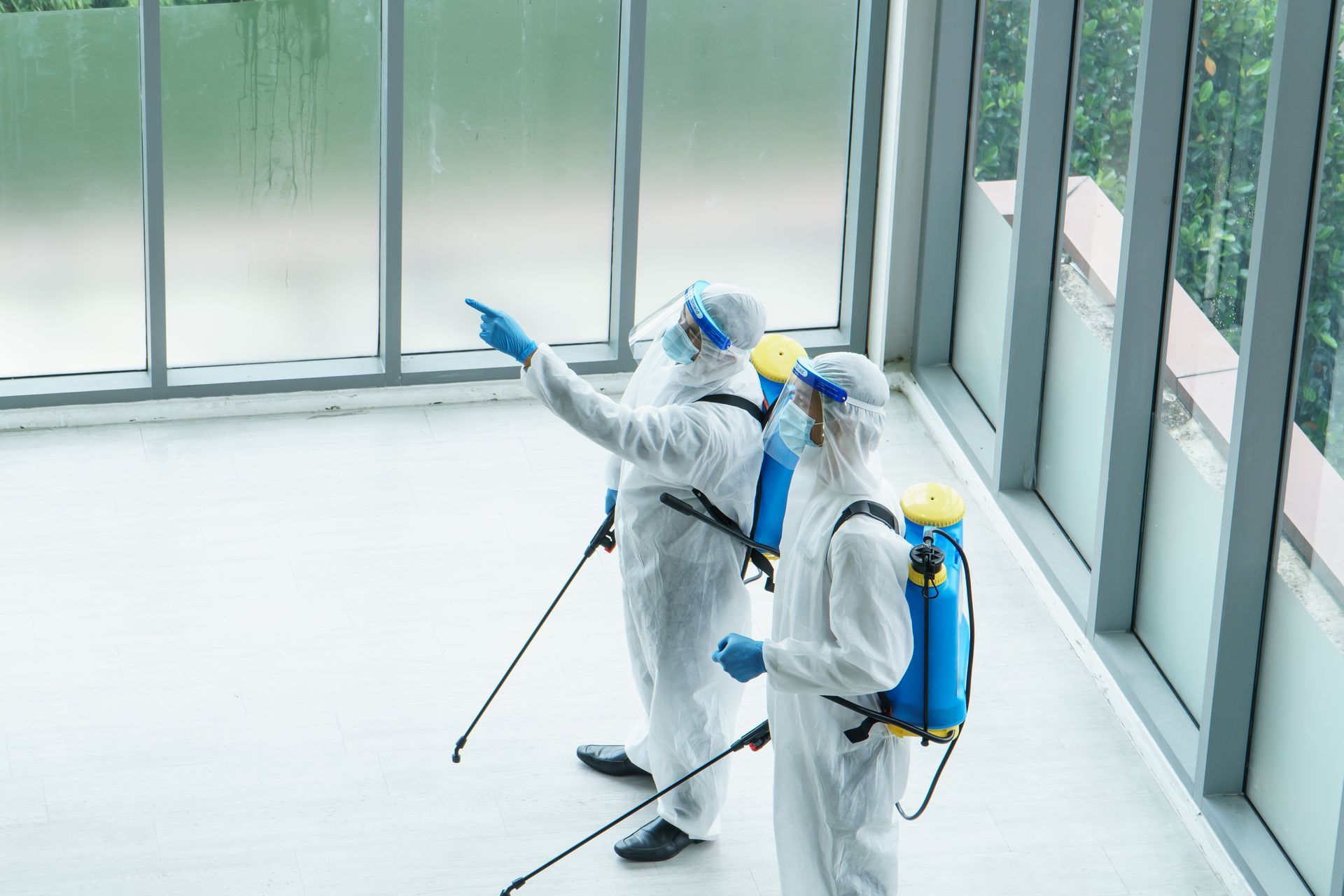 Two workers in full white protective gear and masks use sprayers to disinfect a room with large glass windows.