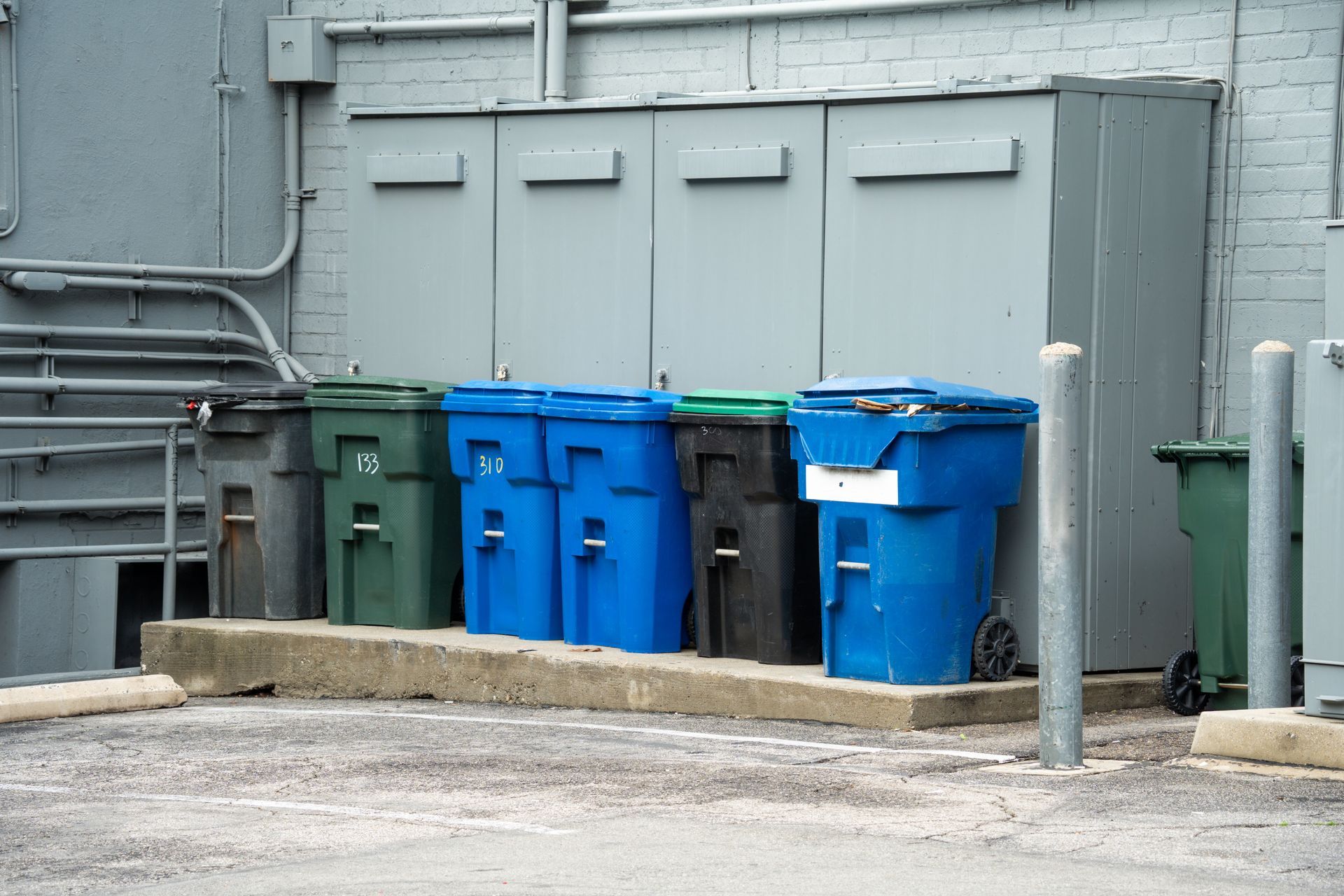 A row of six colorful waste and recycling bins sitting on a concrete ledge against a gray brick wall.
