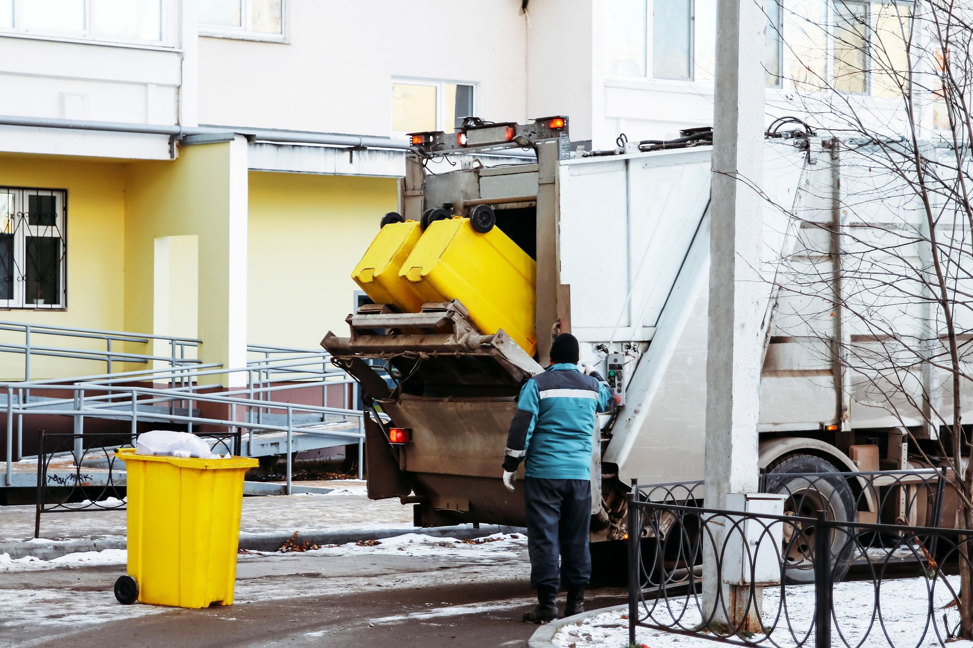 A worker in a turquoise jacket operates a garbage truck, loading bright yellow bins near a building on a winter day.