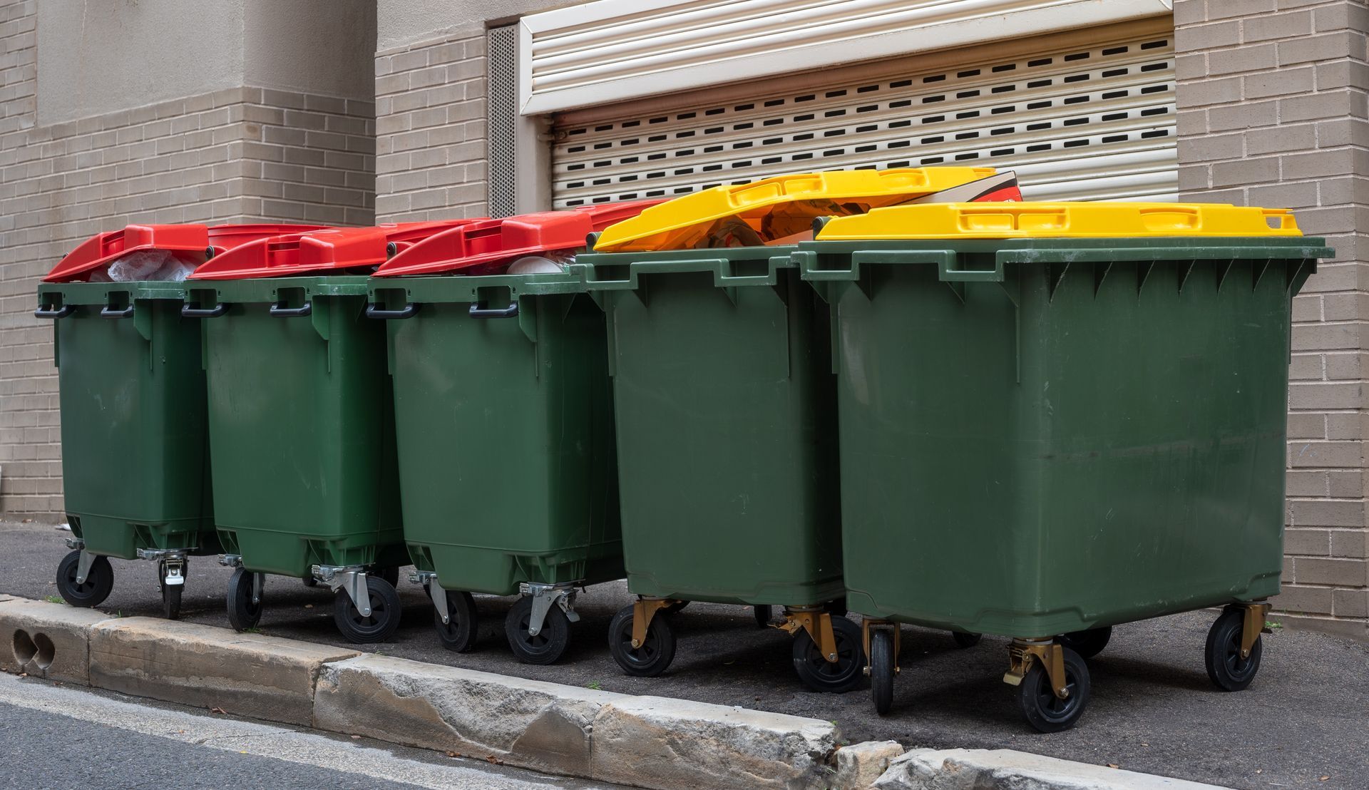 A row of green industrial dumpsters against a brick wall, three with red lids and two with yellow lids.