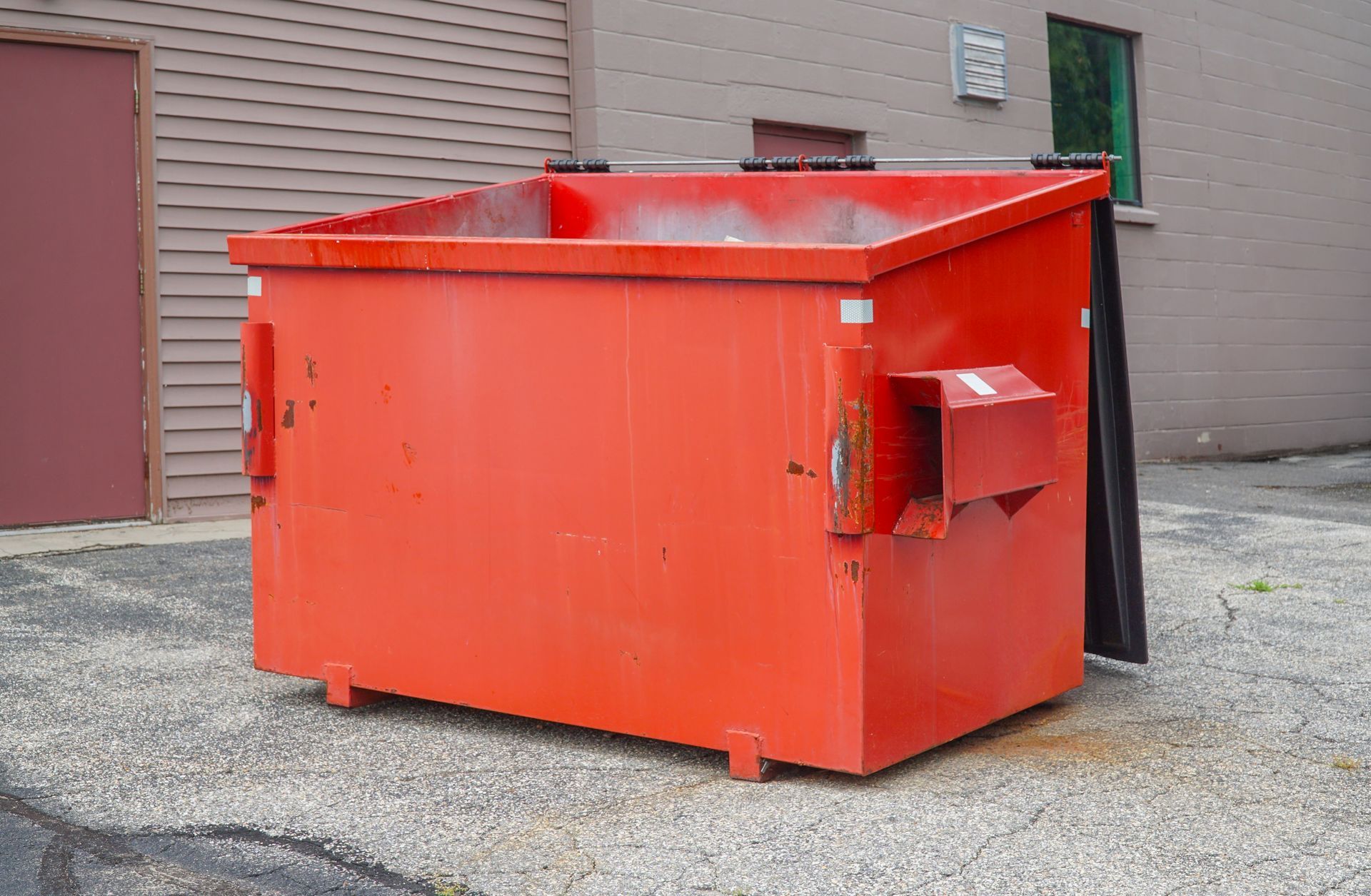 A red, metal dumpster sits on an asphalt surface against the side of a building.
