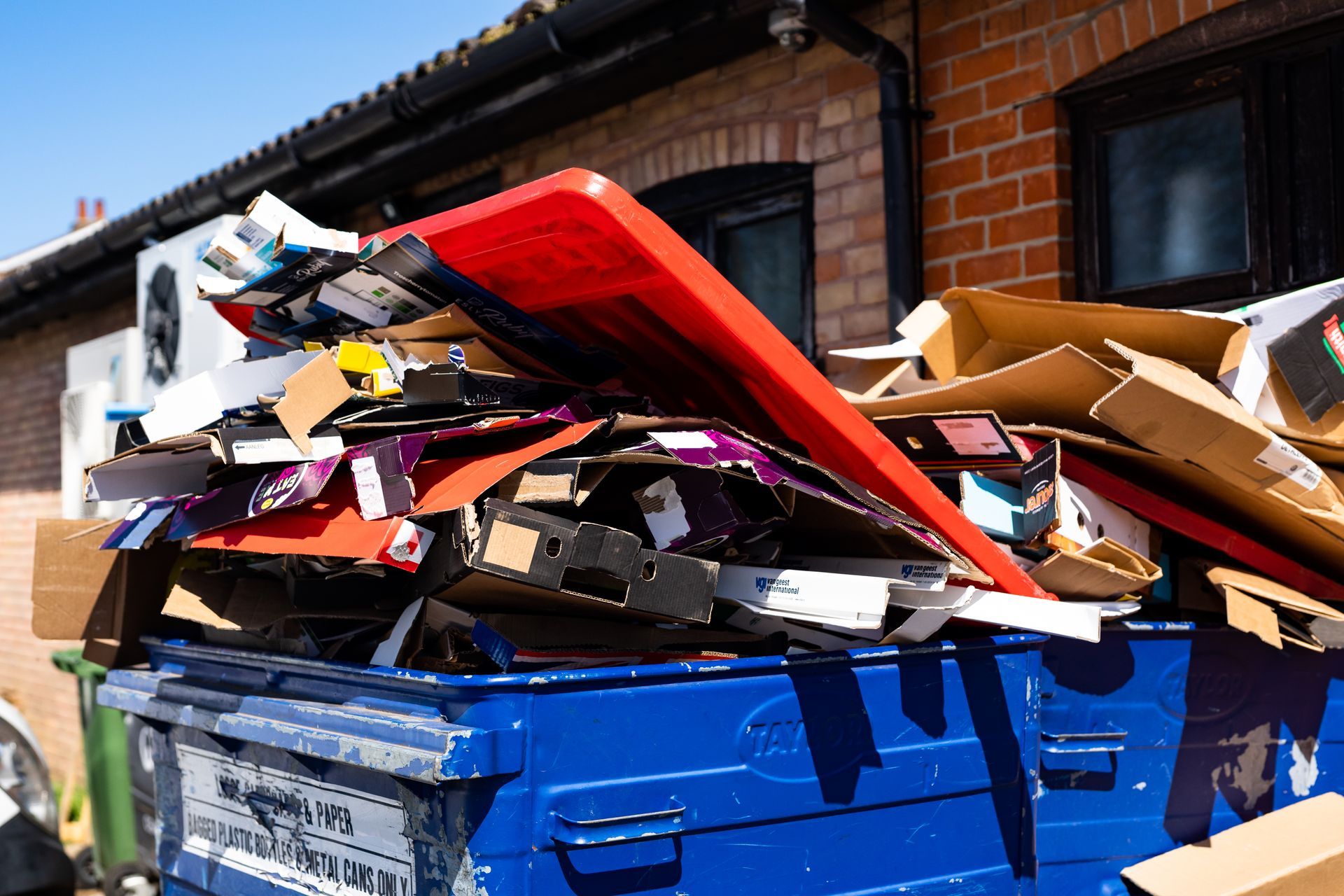 A blue industrial dumpster overflowing with cardboard boxes and a large red plastic board against a brick building.
