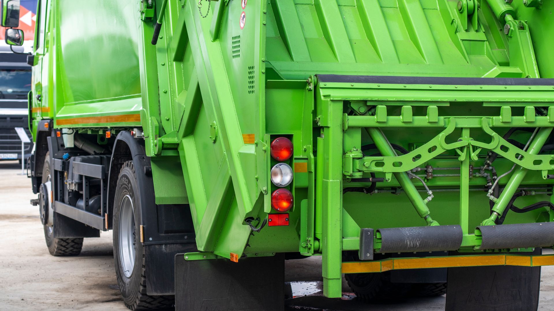 A bright green commercial garbage truck parked on a paved lot, focusing on the rear loading mechanism and taillights.