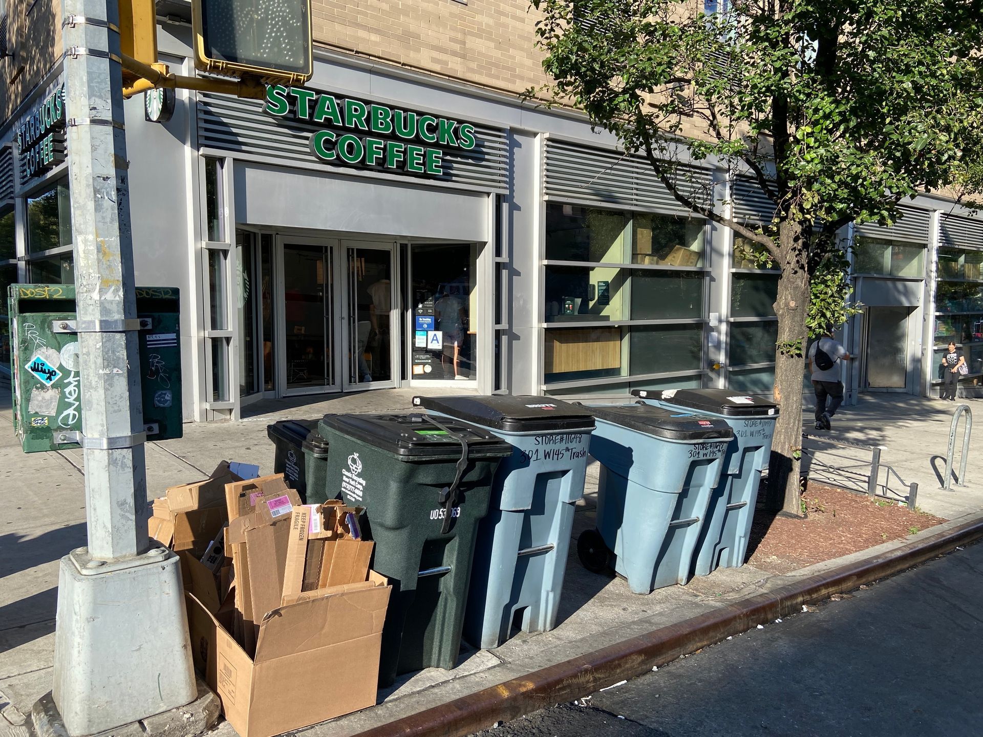 A Starbucks storefront with several dark grey trash bins and a cardboard box on the sidewalk in front of it.