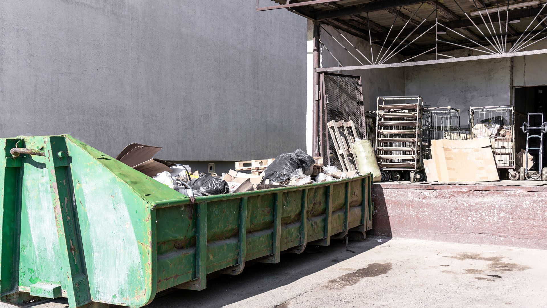 A large green dumpster sits on a concrete pad outside a building with an open loading dock area.