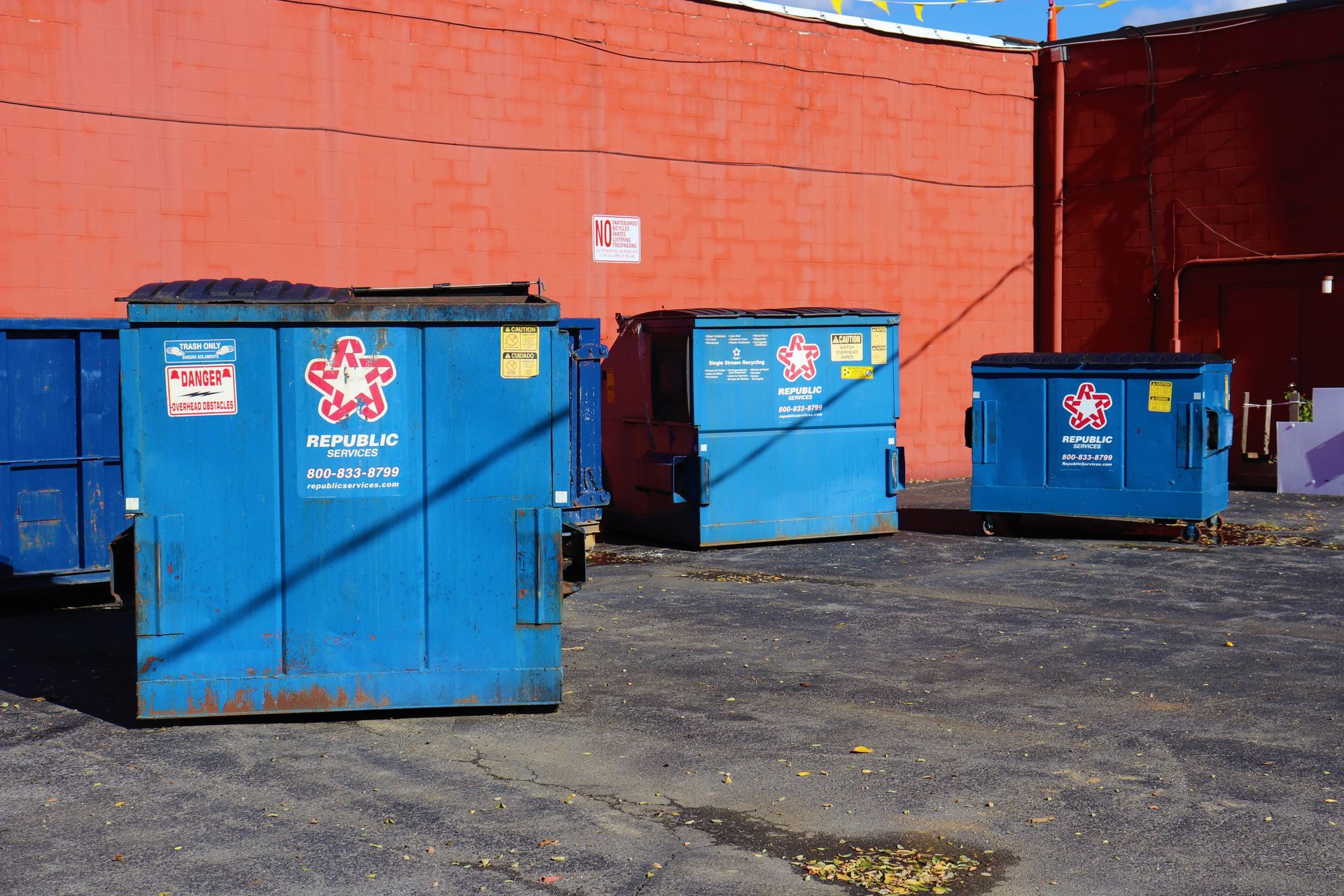 Three blue Republic Services dumpsters sit in a row against a large, bright orange exterior brick wall.