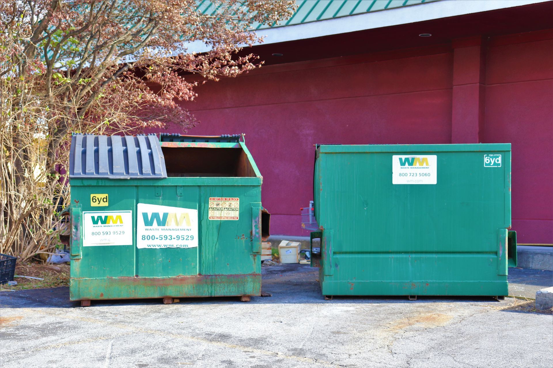Two green Waste Management dumpsters sitting on asphalt in front of a red building wall and a tree.