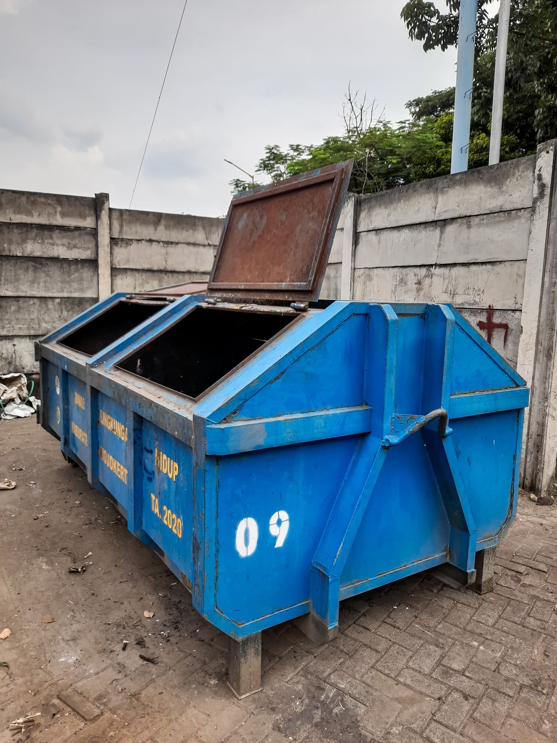 A blue, rectangular industrial dumpster with an open lid, marked with the number 09, sitting on a paved surface.