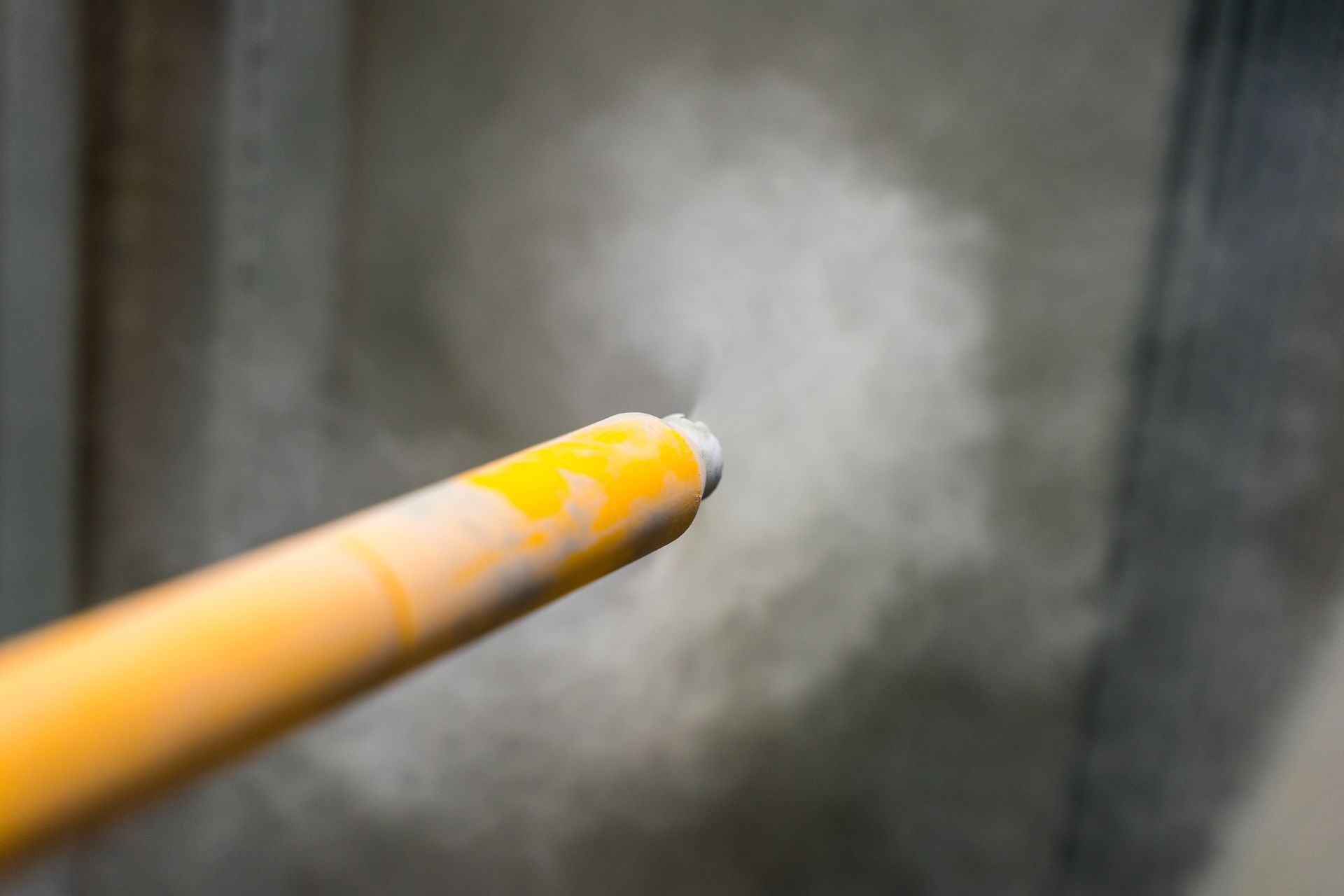 A yellow industrial nozzle sprays a fine mist or smoke against a blurred, gray background.