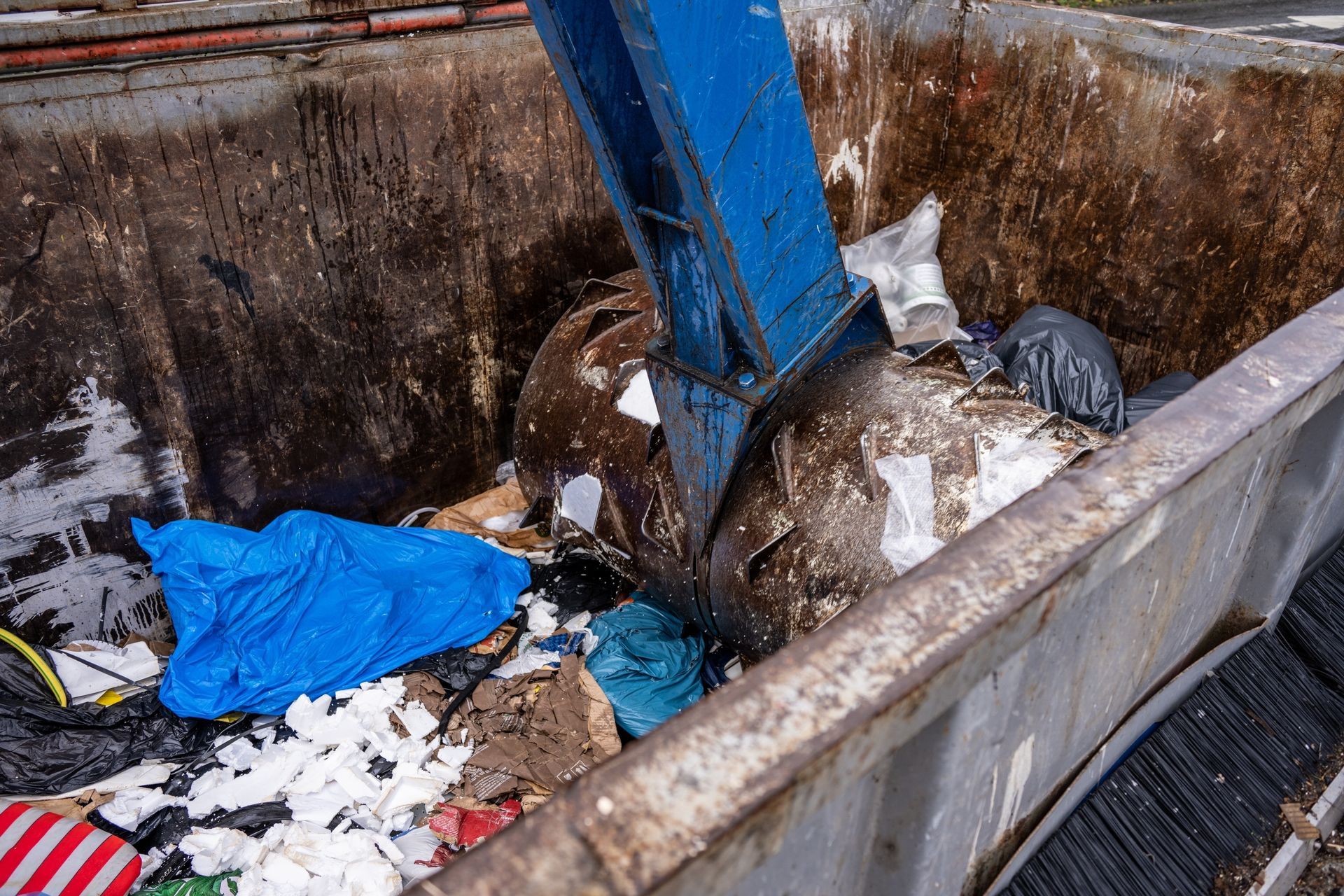 A blue industrial claw picks up a large, rusted metal cylinder from inside a dumpster filled with debris and trash.