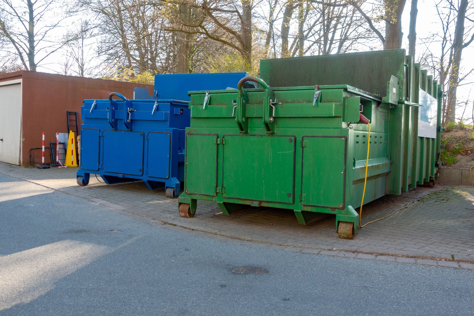 A blue and a large green industrial waste container sit parked on a paved lot beside a building.