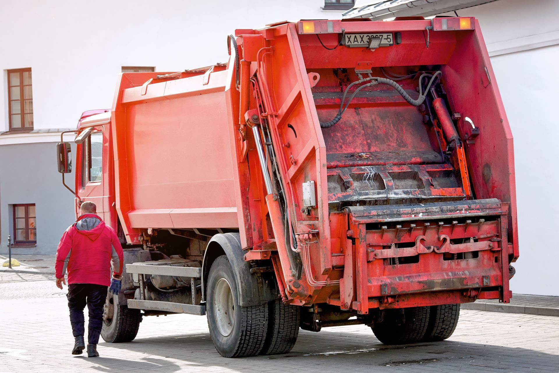 A person in a red jacket walks toward a bright red garbage truck parked on a paved street.