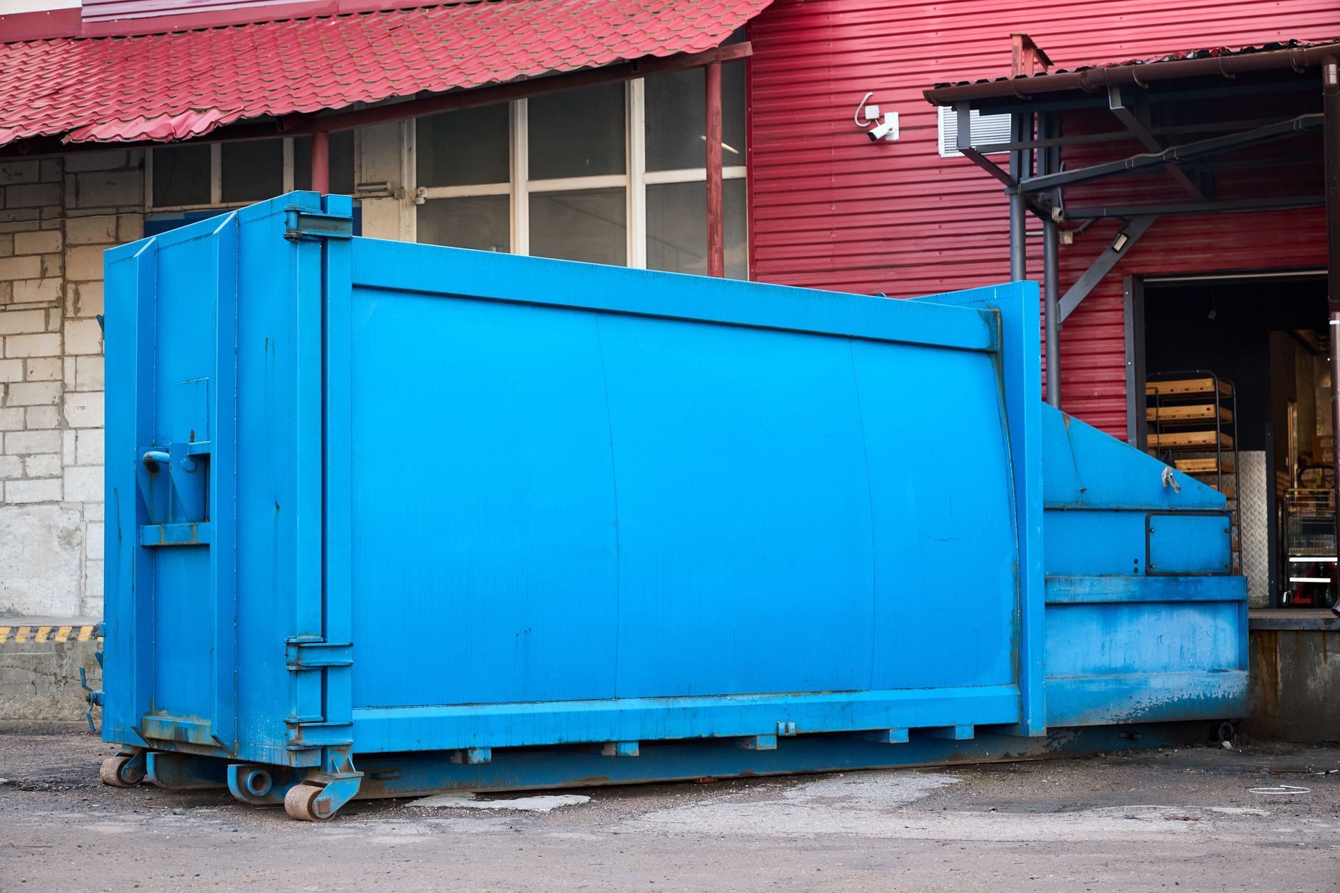 A large blue industrial waste container sits on a concrete lot in front of a red building.