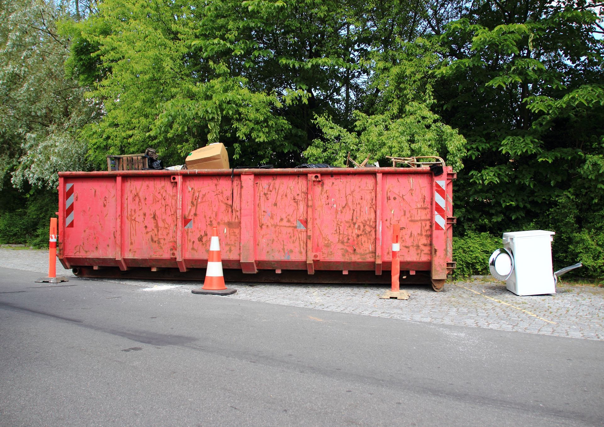 A large red industrial dumpster sits on a gravel shoulder next to an asphalt road, with traffic cones and a white appliance.