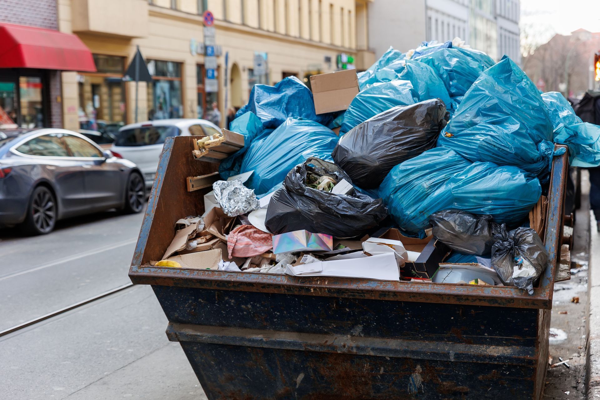 An overflowing metal dumpster on a city street, filled with blue and black plastic trash bags and loose cardboard.