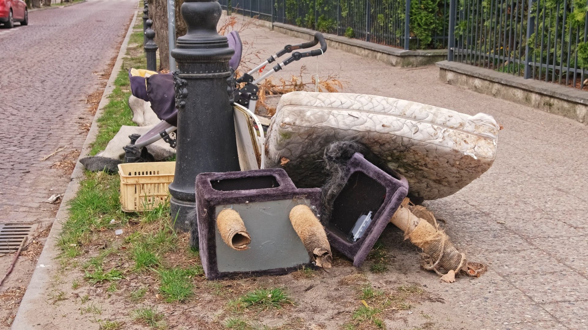 A pile of discarded household items, including a mattress and a cat tower, sitting on a sidewalk next to a street lamp.