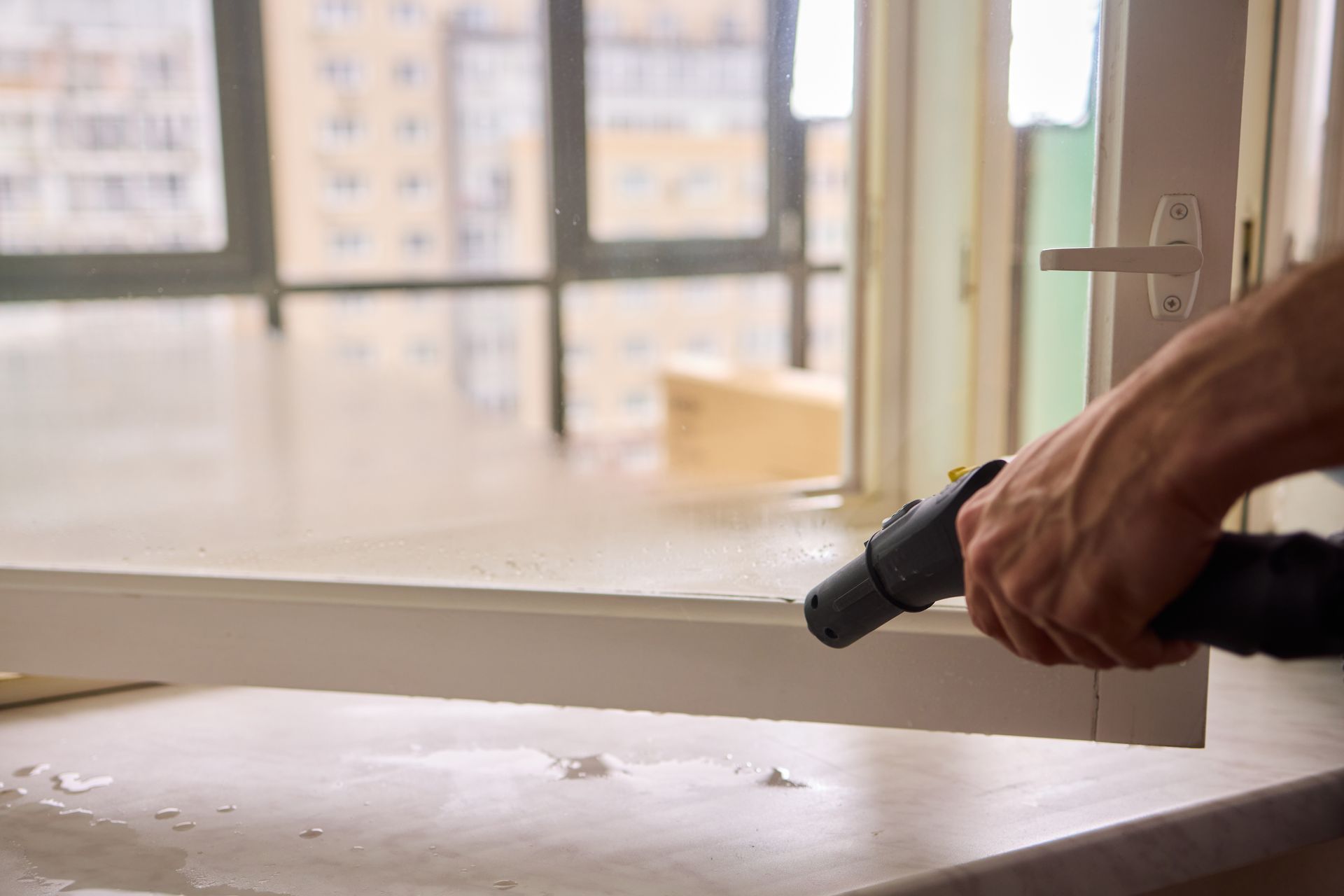 A hand holds a steam cleaner nozzle, cleaning the white frame of a window in a room with a view of other buildings.