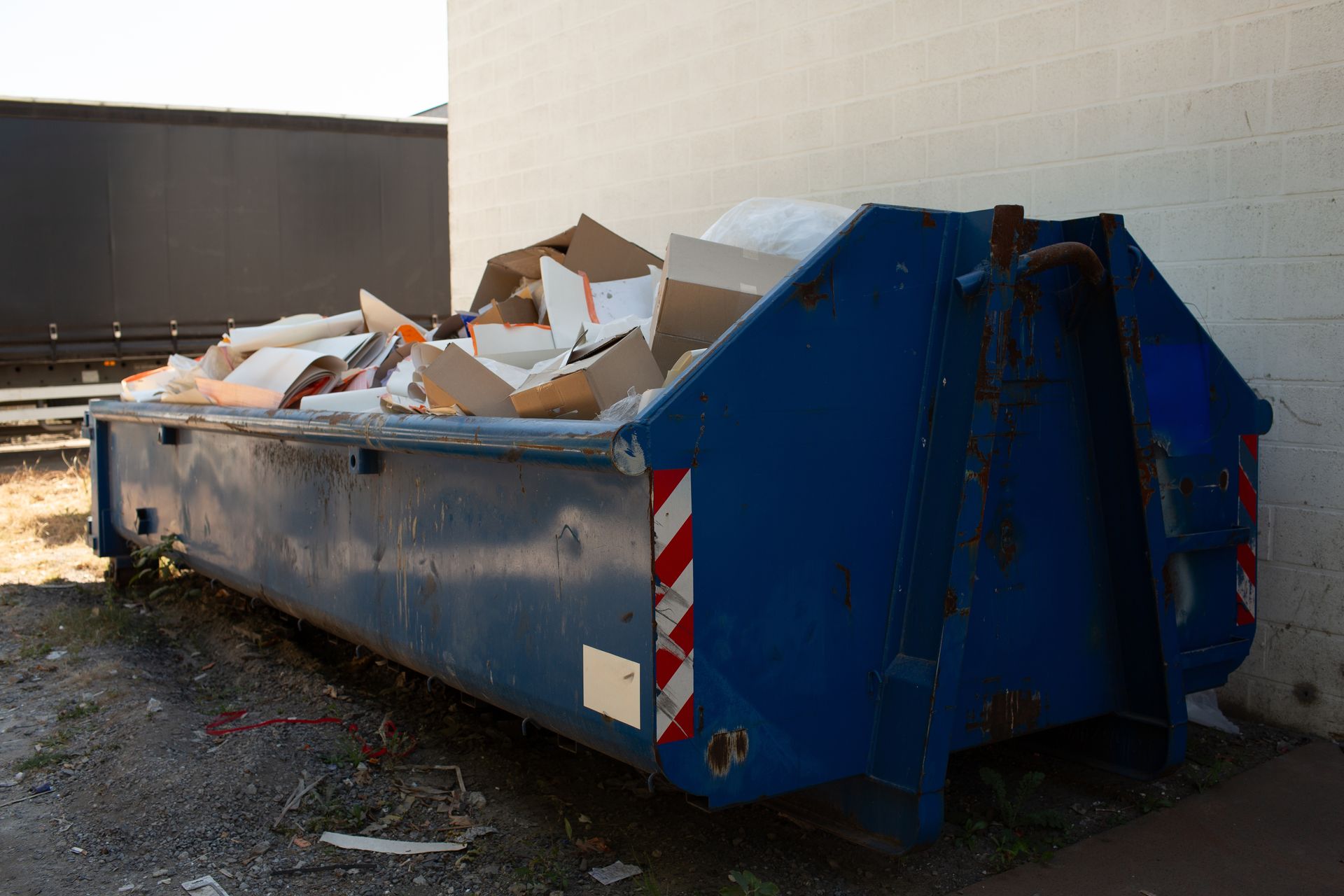 A large, blue metal dumpster filled with cardboard boxes sits against a white brick wall.