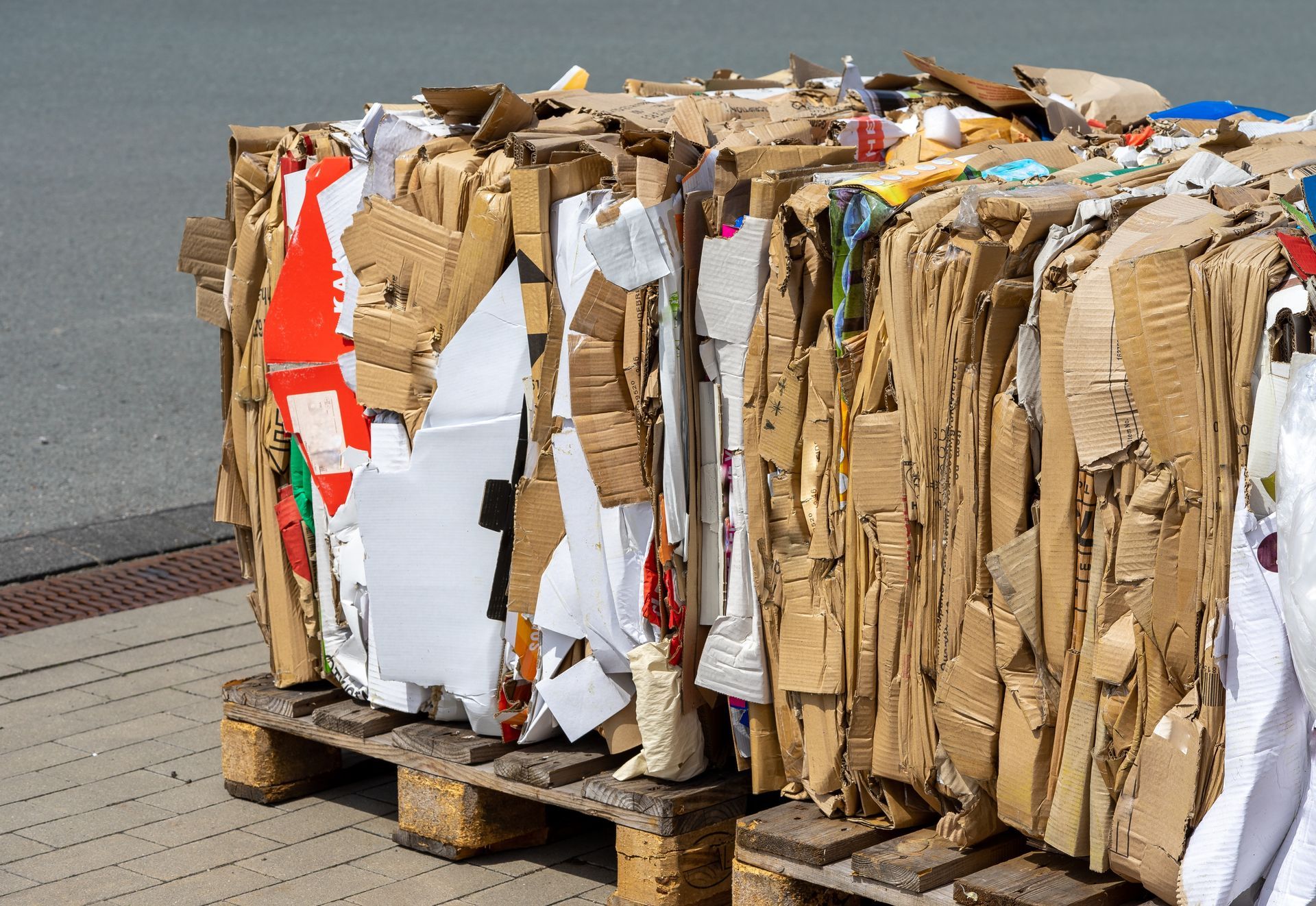 A large, rectangular bale of compressed cardboard and paper waste sits on a wooden pallet outdoors on a paved surface.