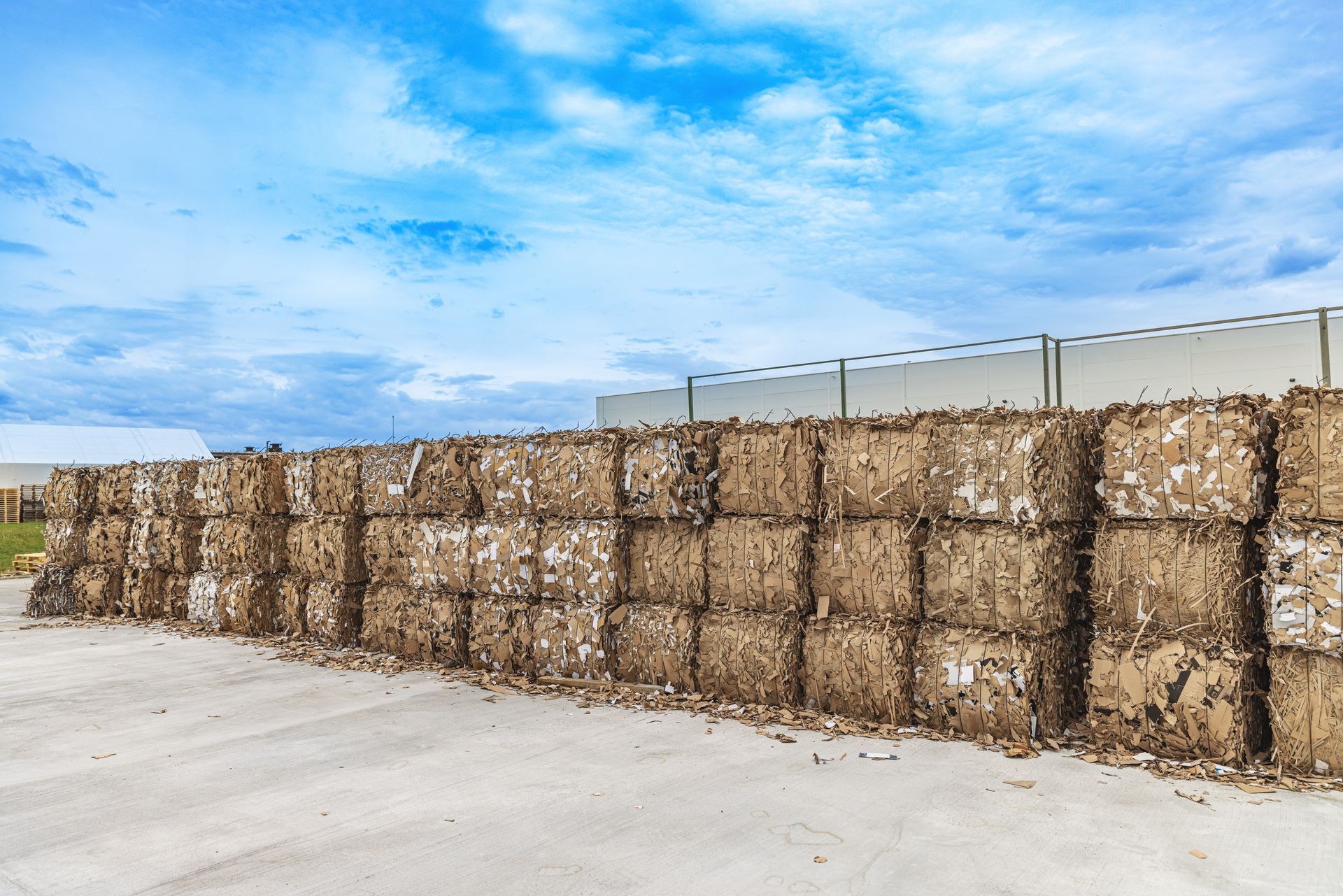 A long wall of neatly stacked, square bales of compressed waste material on a concrete lot under a blue, cloudy sky.