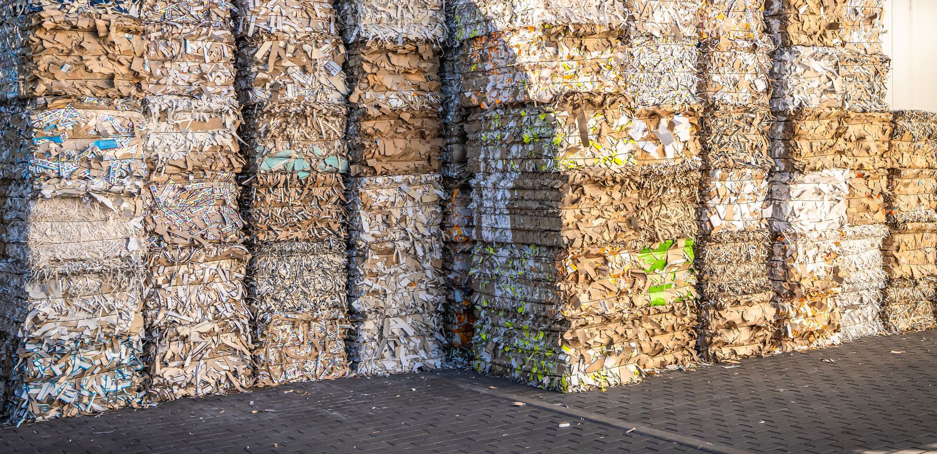 A large stack of compressed, rectangular paper and cardboard bales stored outside on a paved lot.
