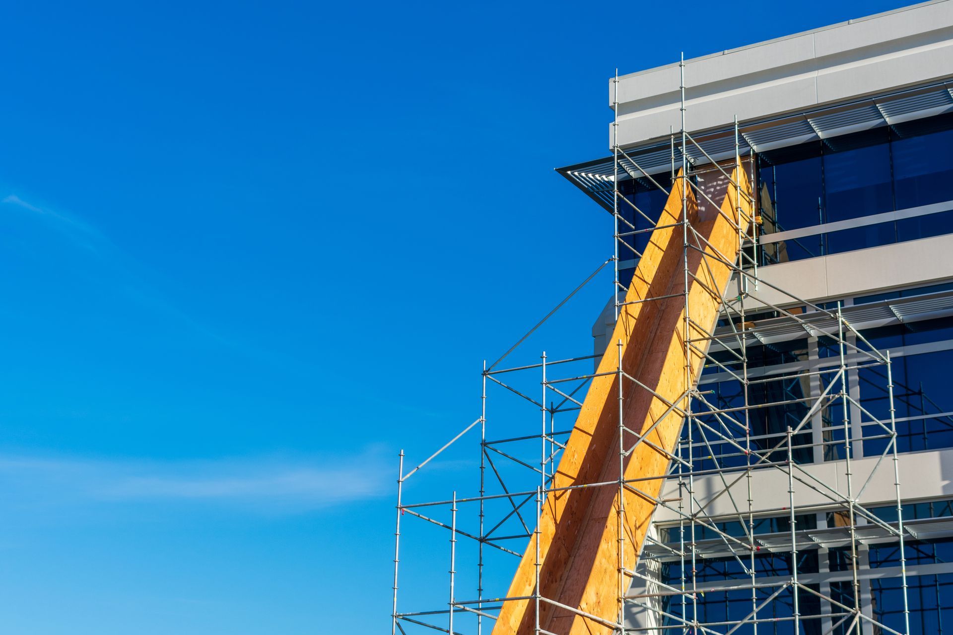 Yellow debris chute attached to scaffolding on the side of a modern glass office building against a blue sky.