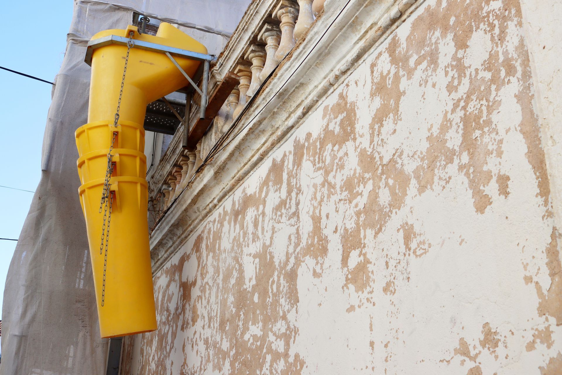 A yellow debris chute mounted to the exterior of a weathered, peeling plaster building.