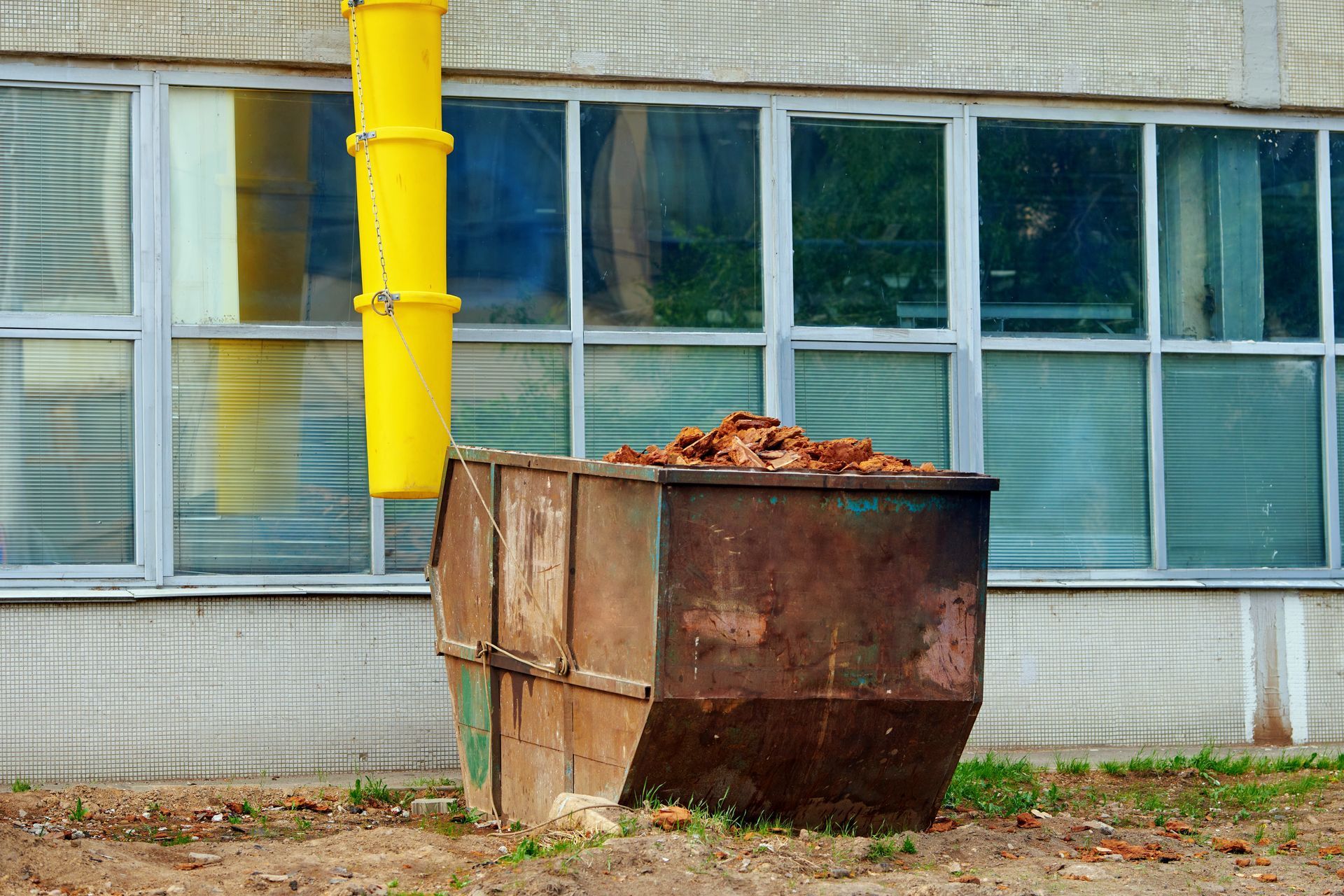 A rusty metal dumpster filled with construction debris sits beneath a yellow plastic debris chute against a building.