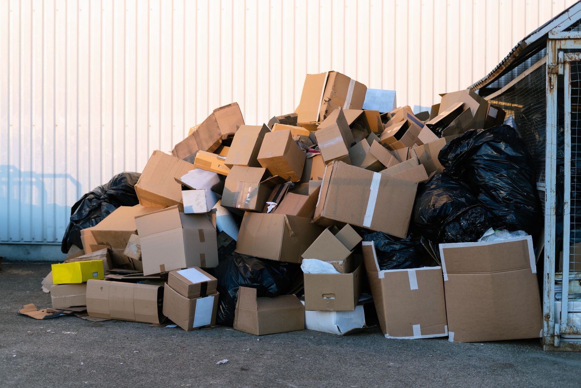 A large, disorganized pile of brown cardboard boxes and black plastic trash bags outdoors against a corrugated wall.
