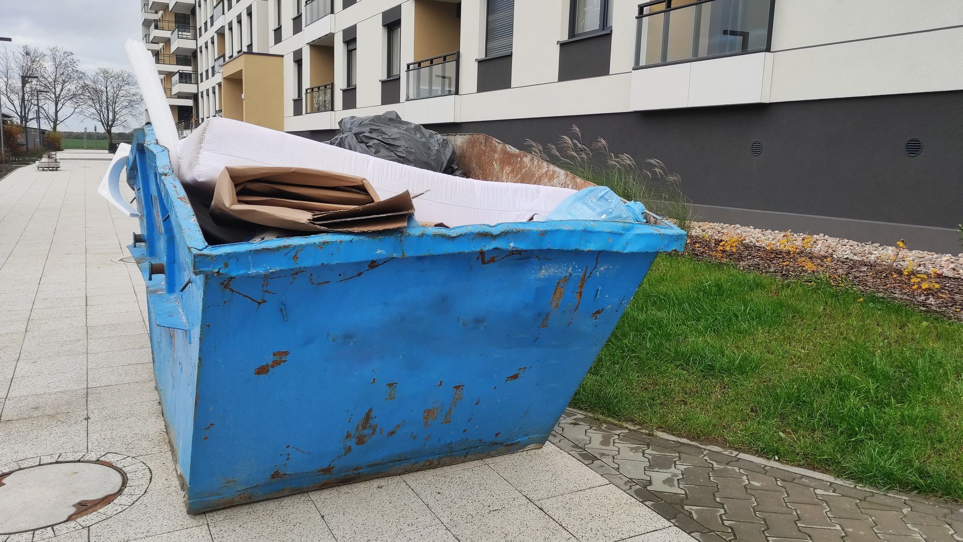 A blue construction dumpster sits on a sidewalk next to a residential building with white walls and a grassy area.