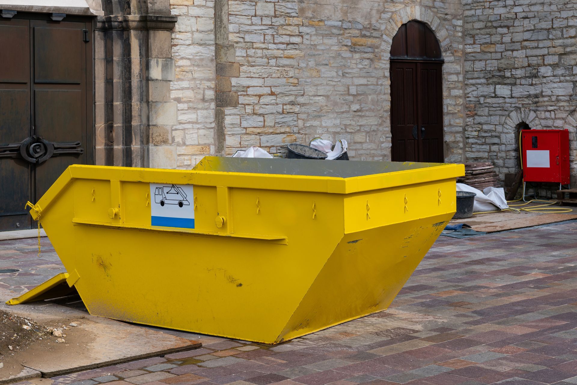 A bright yellow metal waste skip sits on a cobblestone plaza in front of an old stone building with arched doorways.