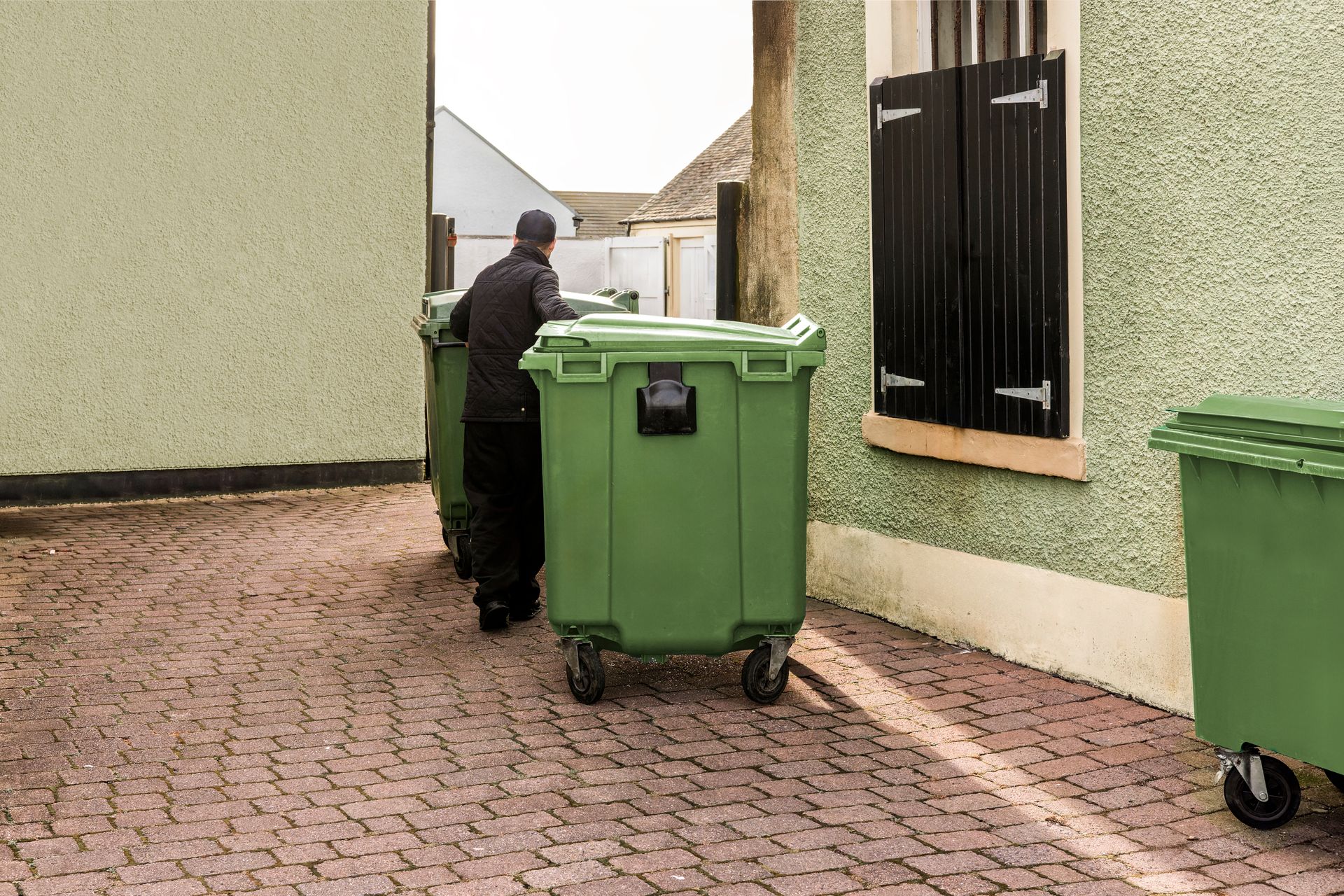 A person wearing dark clothing moves a large, green rolling waste bin along a stone-paved alleyway.