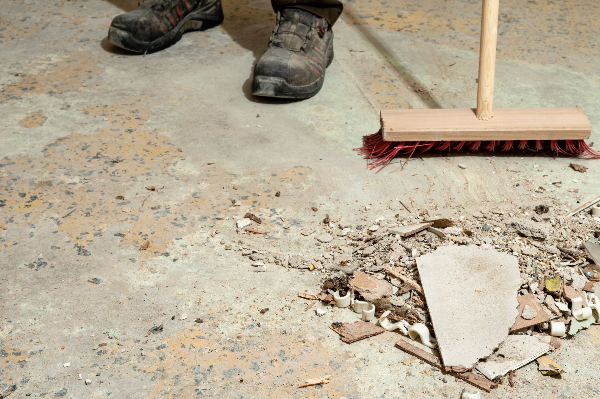A person in work boots uses a push broom to sweep debris off a concrete floor.