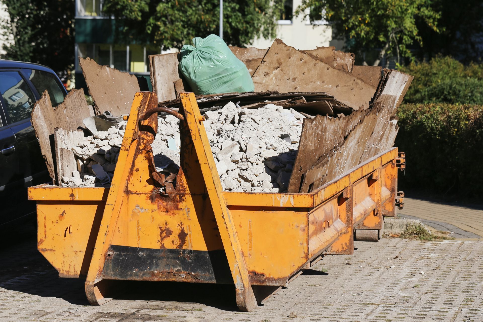 A yellow construction dumpster filled with building debris, pieces of wood, and a green bag on a paved surface.