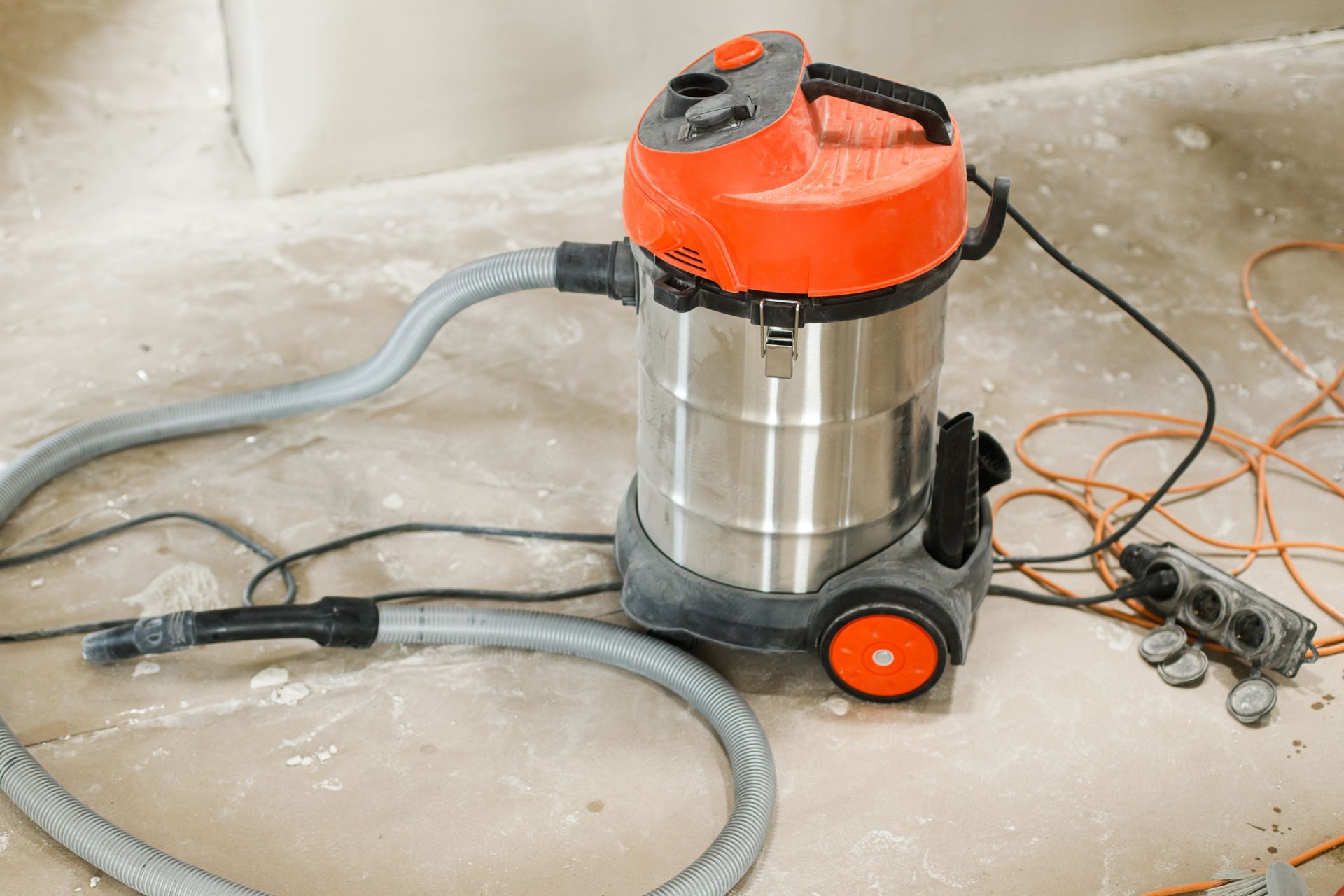 An orange and silver industrial shop vacuum stands on a concrete floor with a hose and power strip nearby.