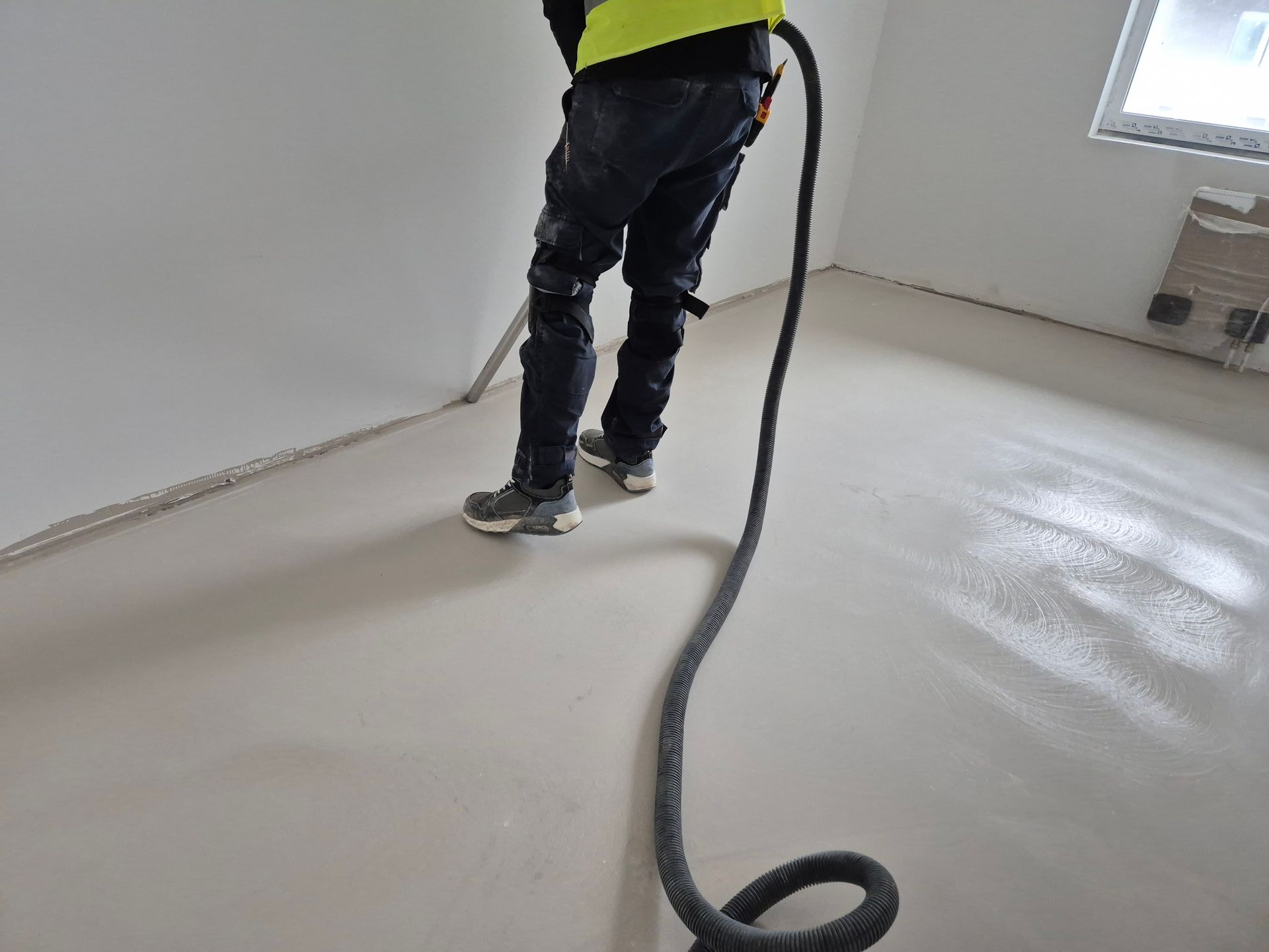 A construction worker in a high-visibility vest uses a long hose to spread self-leveling floor compound in a room.