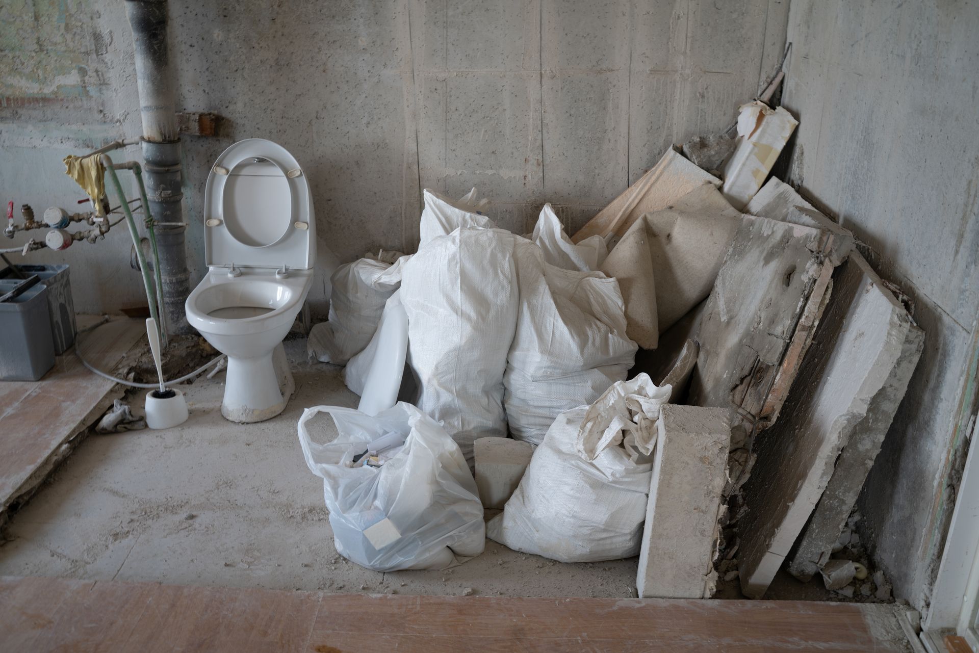 A toilet sits in an unfinished room next to a pile of white debris bags and construction materials against a gray wall.