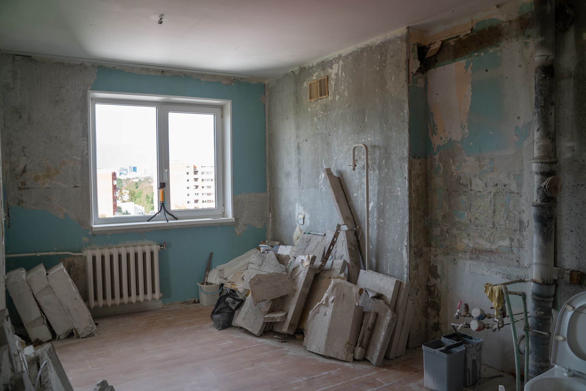 A room under renovation with bare walls, a white radiator, a window, and a large pile of construction debris.