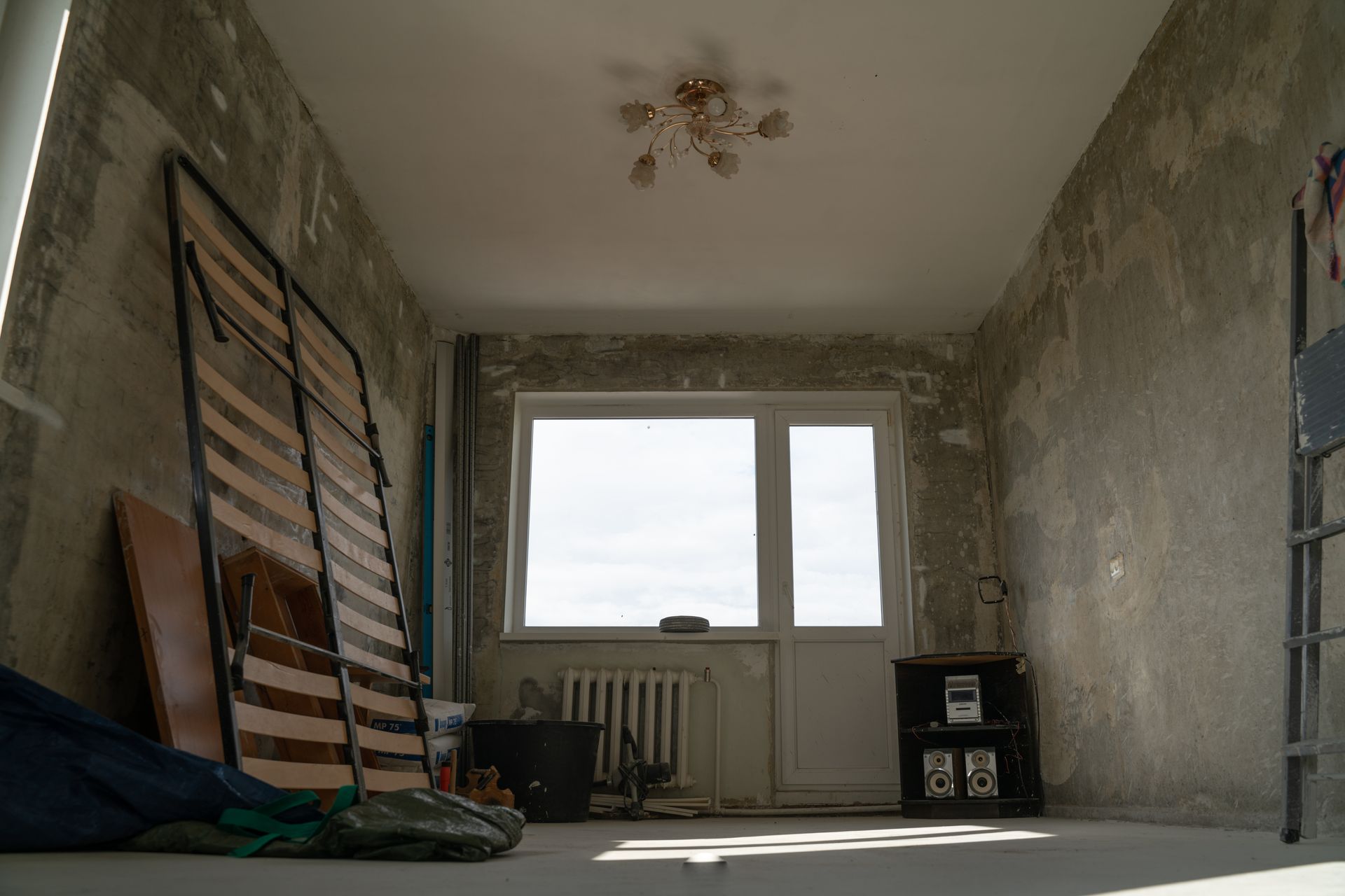 A sparsely furnished room undergoing renovation with unfinished gray walls, a wooden bed frame, and a window with a door.