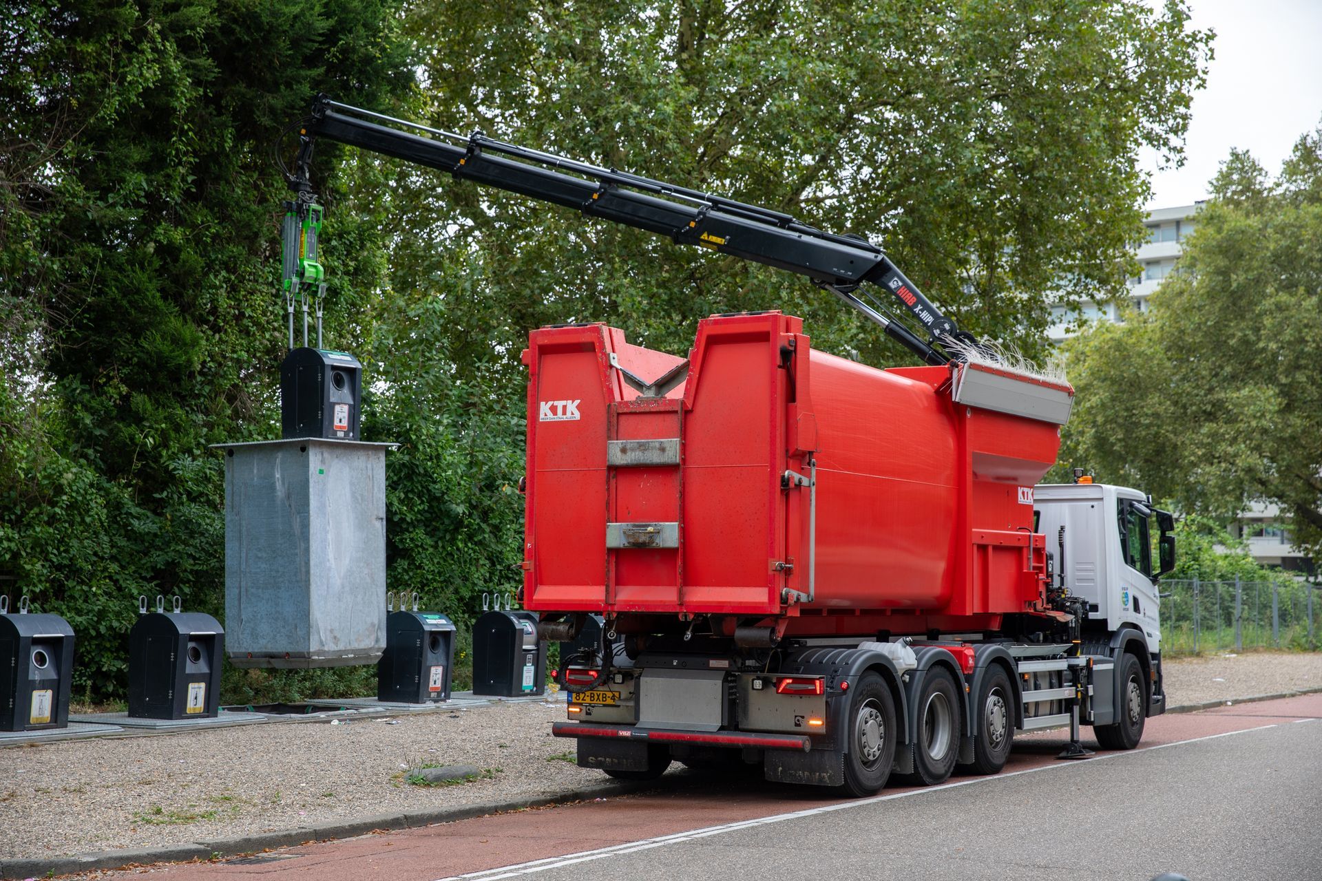 A red crane truck lifts a grey recycling container from a row of bins beside a road.