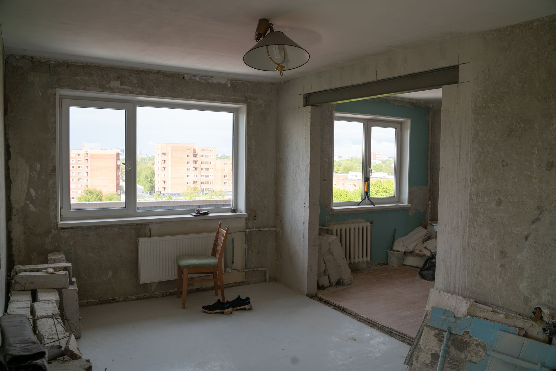 An empty room undergoing renovation with exposed concrete walls, two large windows, a wooden chair, and debris.