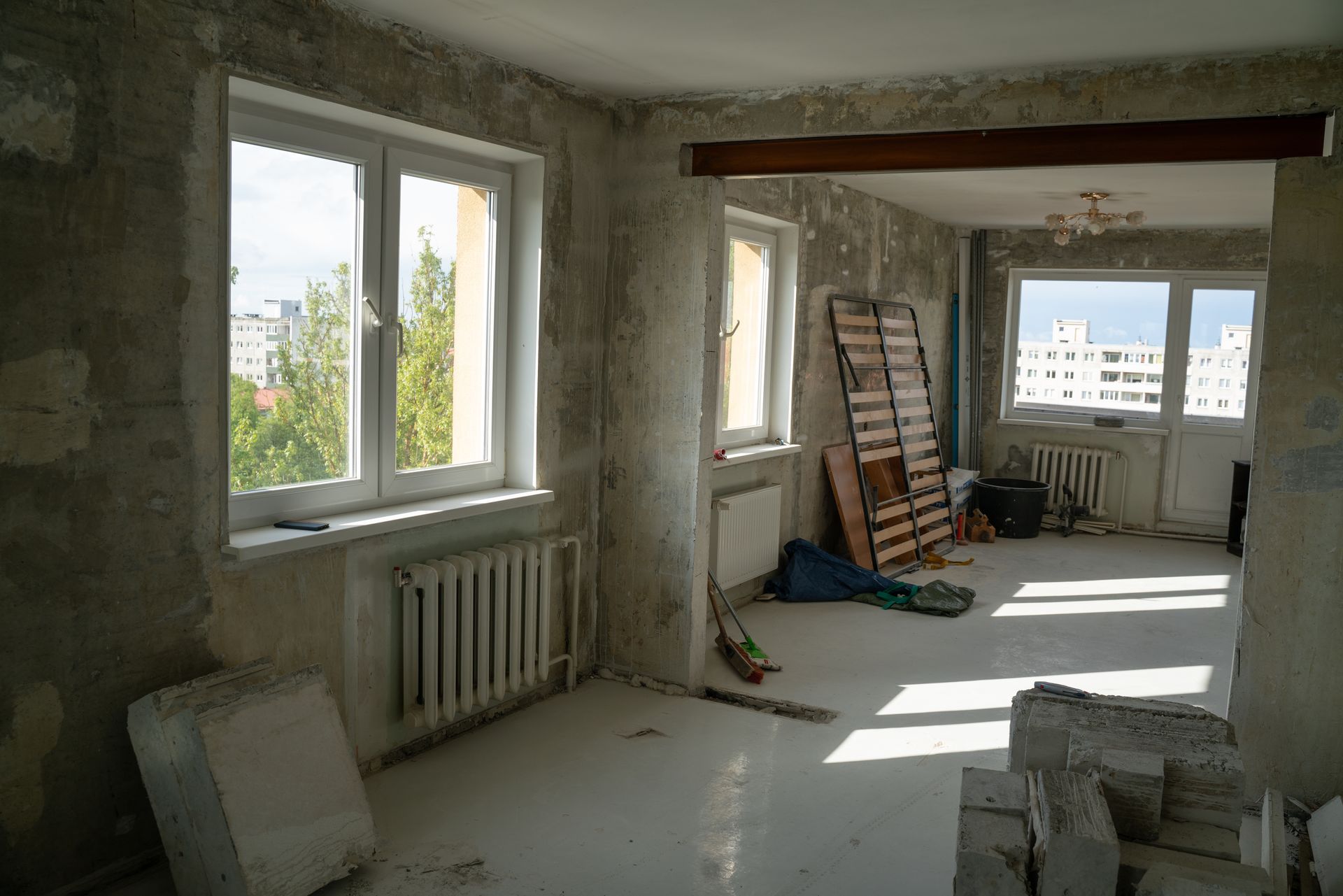 An apartment interior under renovation with bare concrete walls, white floors, windows, and construction debris.