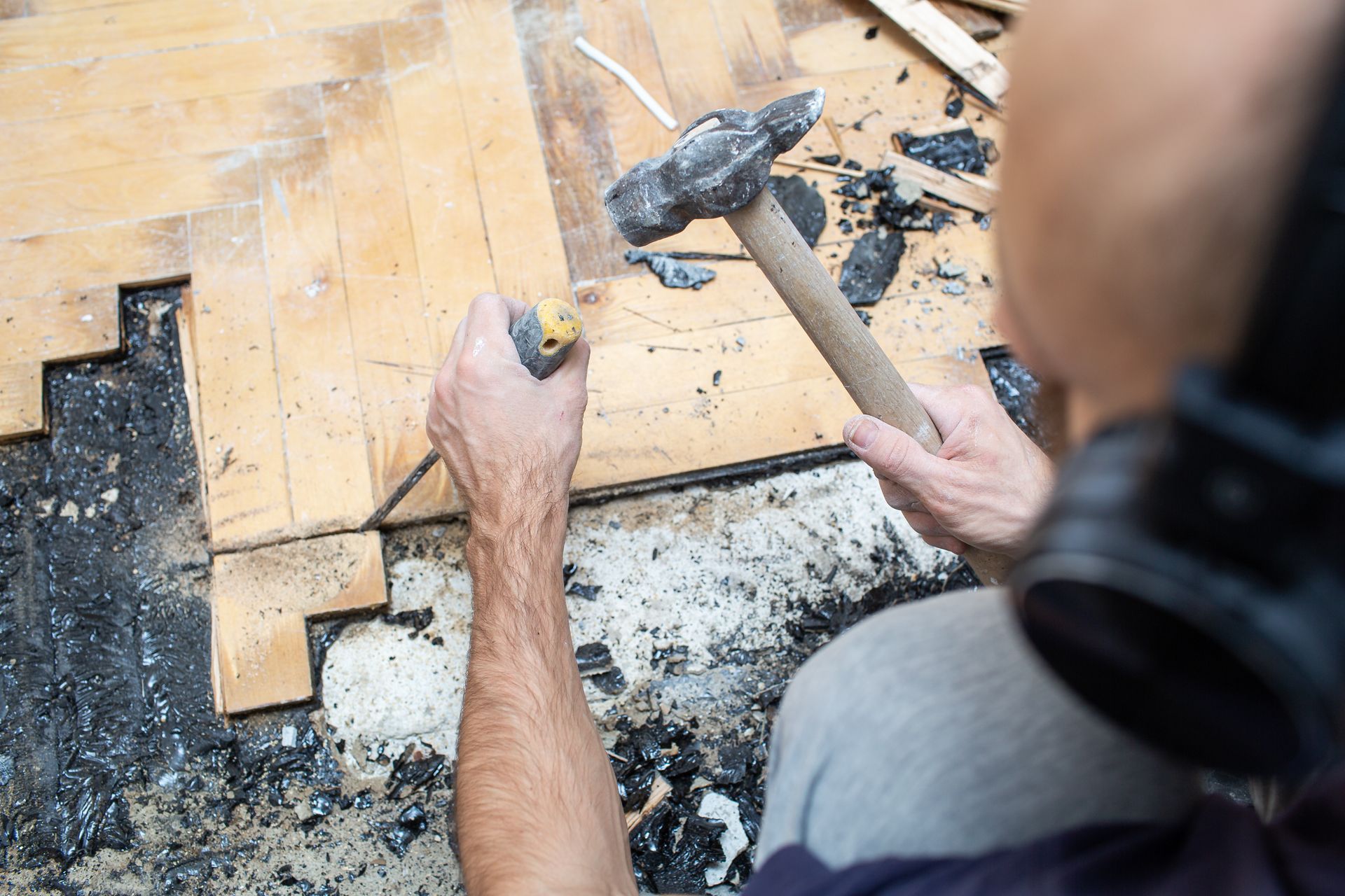 A person wears headphones while using a hammer and chisel to remove old wooden floorboards.