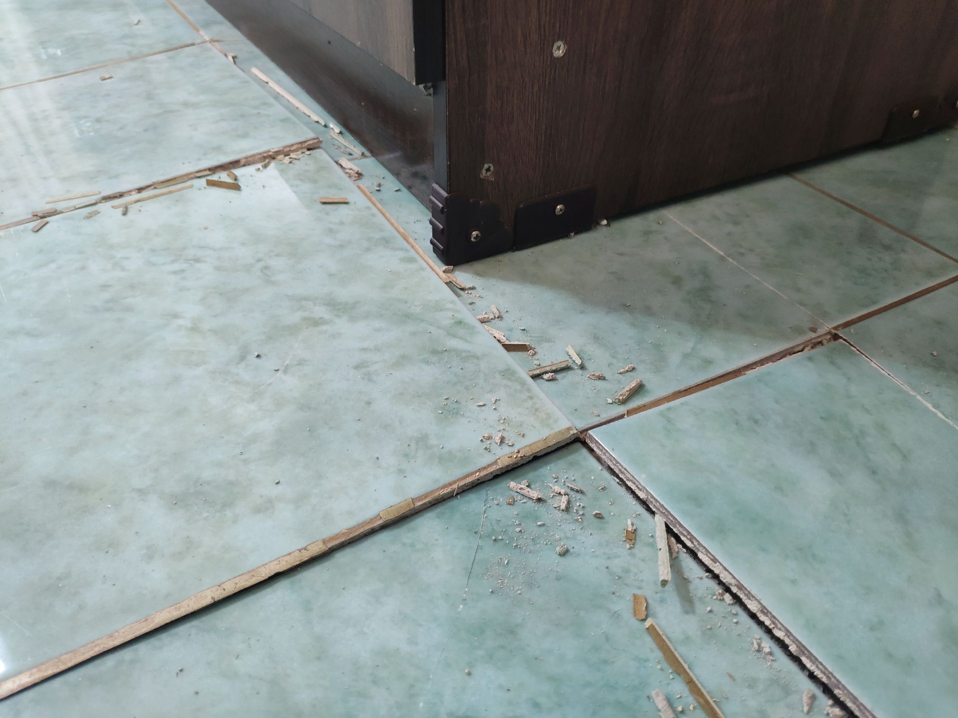 Debris and dust scattered on green tiled flooring near the corner of a dark wooden cabinet.
